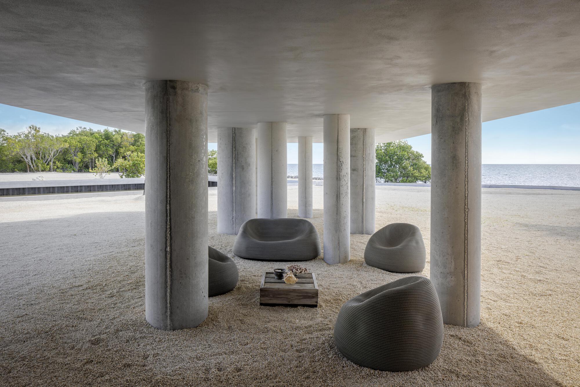 Shaded ground-floor seating area beneath cylindrical concrete columns with gravel floor and coastal view