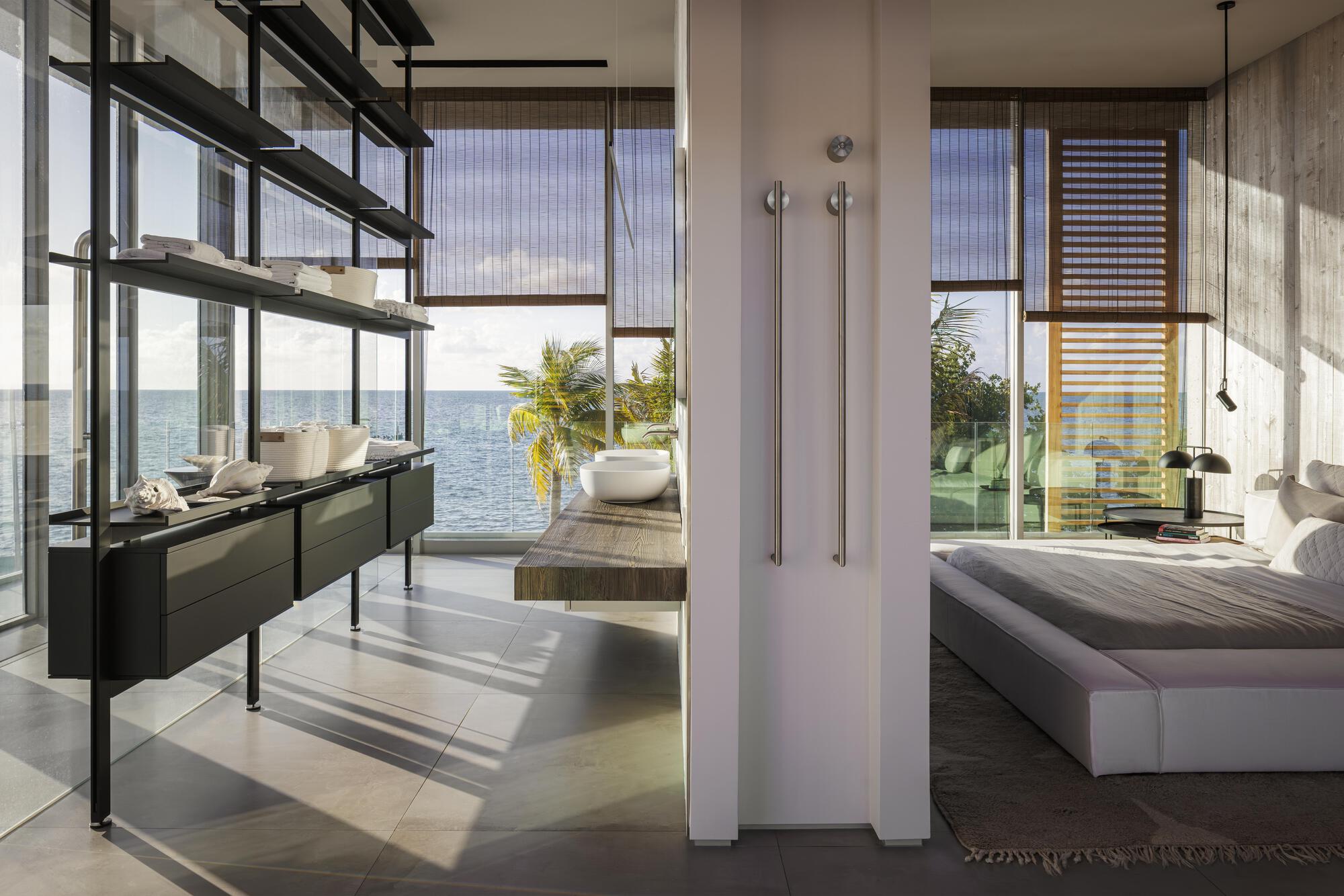 Open bedroom suite with freestanding white partition and full-height glazing framing coastal vegetation