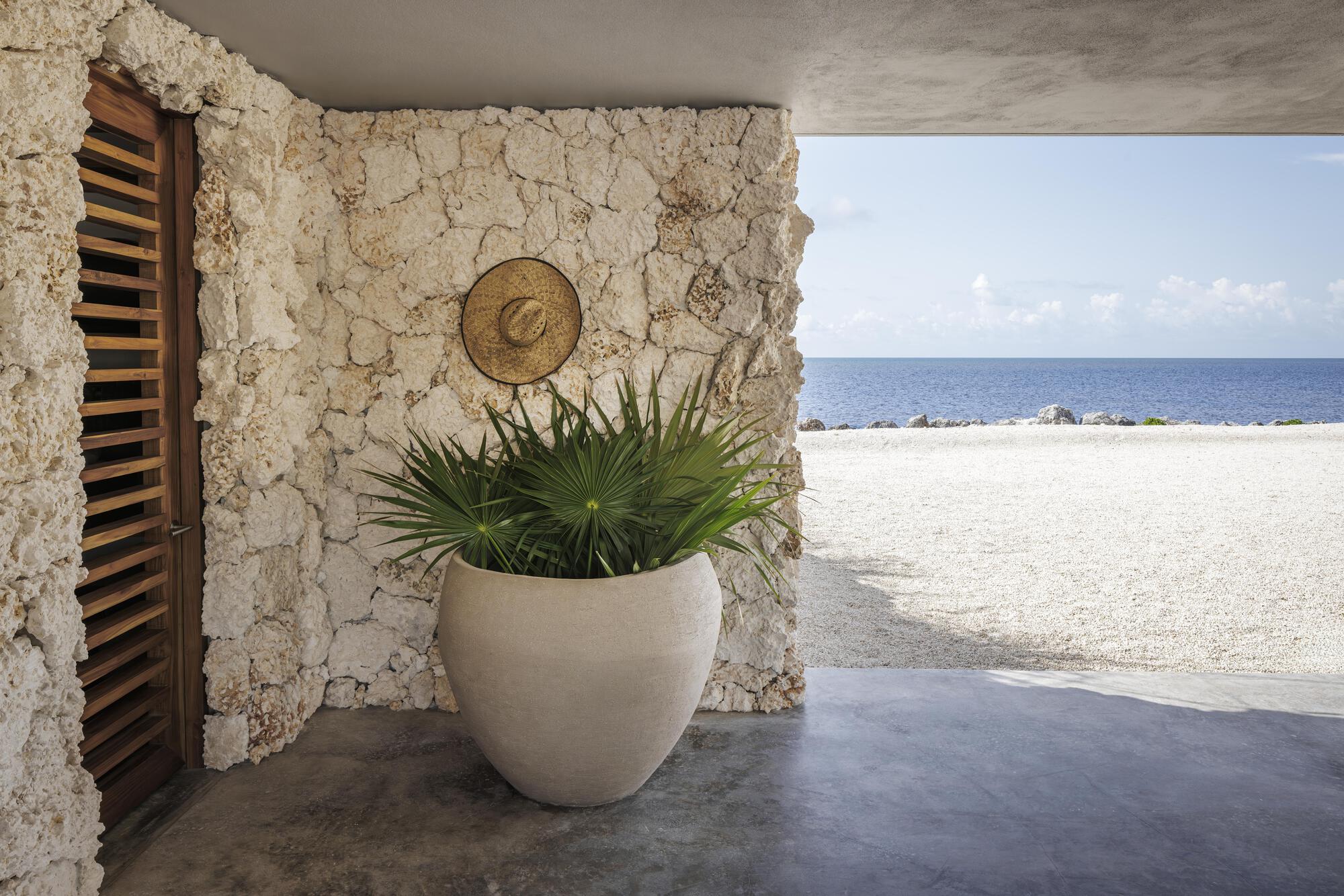 Shaded porch corner with coral stone wall, timber louver door, planted urn, and view to white gravel and ocean