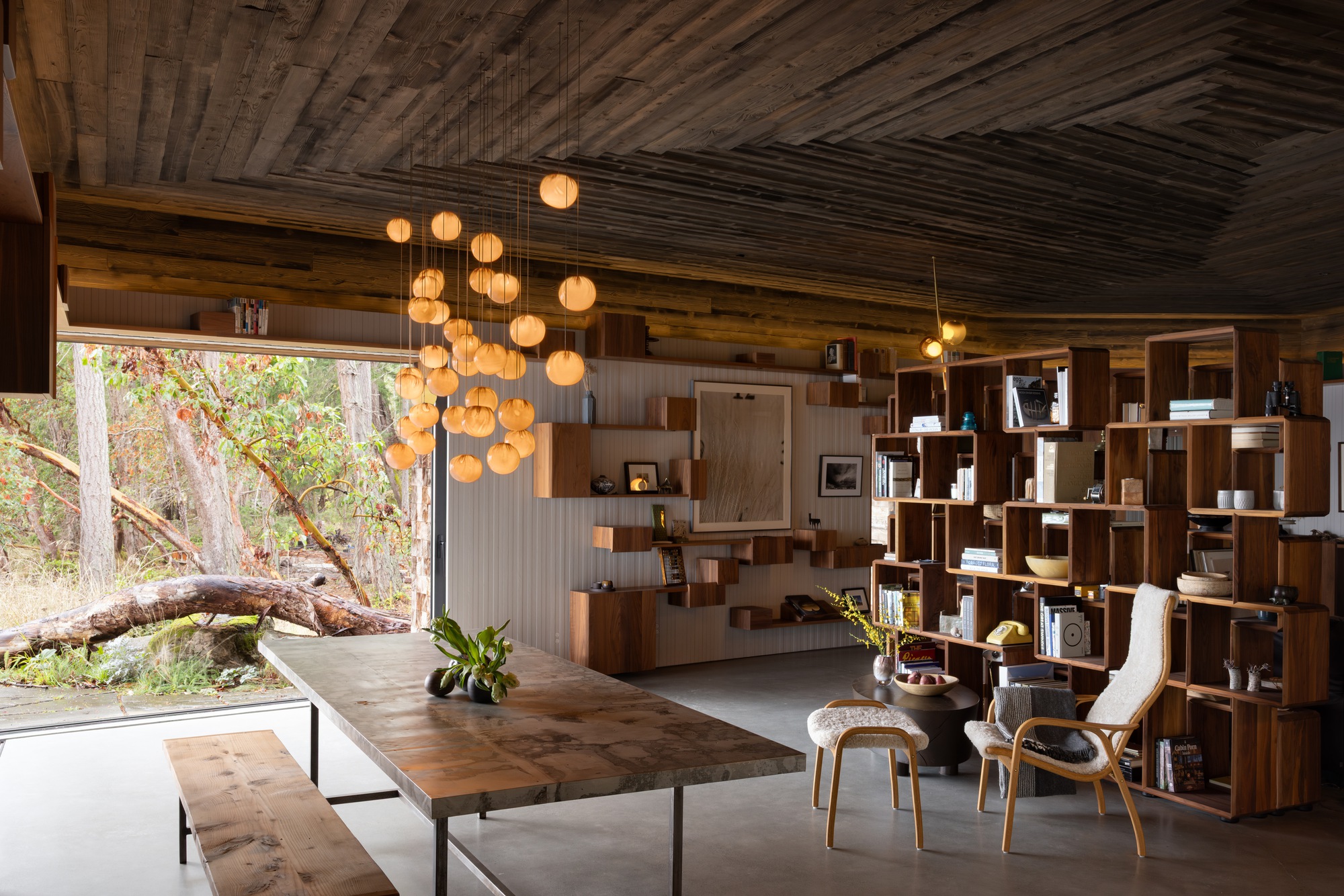 Open living space with wood ceiling, modular shelving wall, and large glazed opening to forest beyond