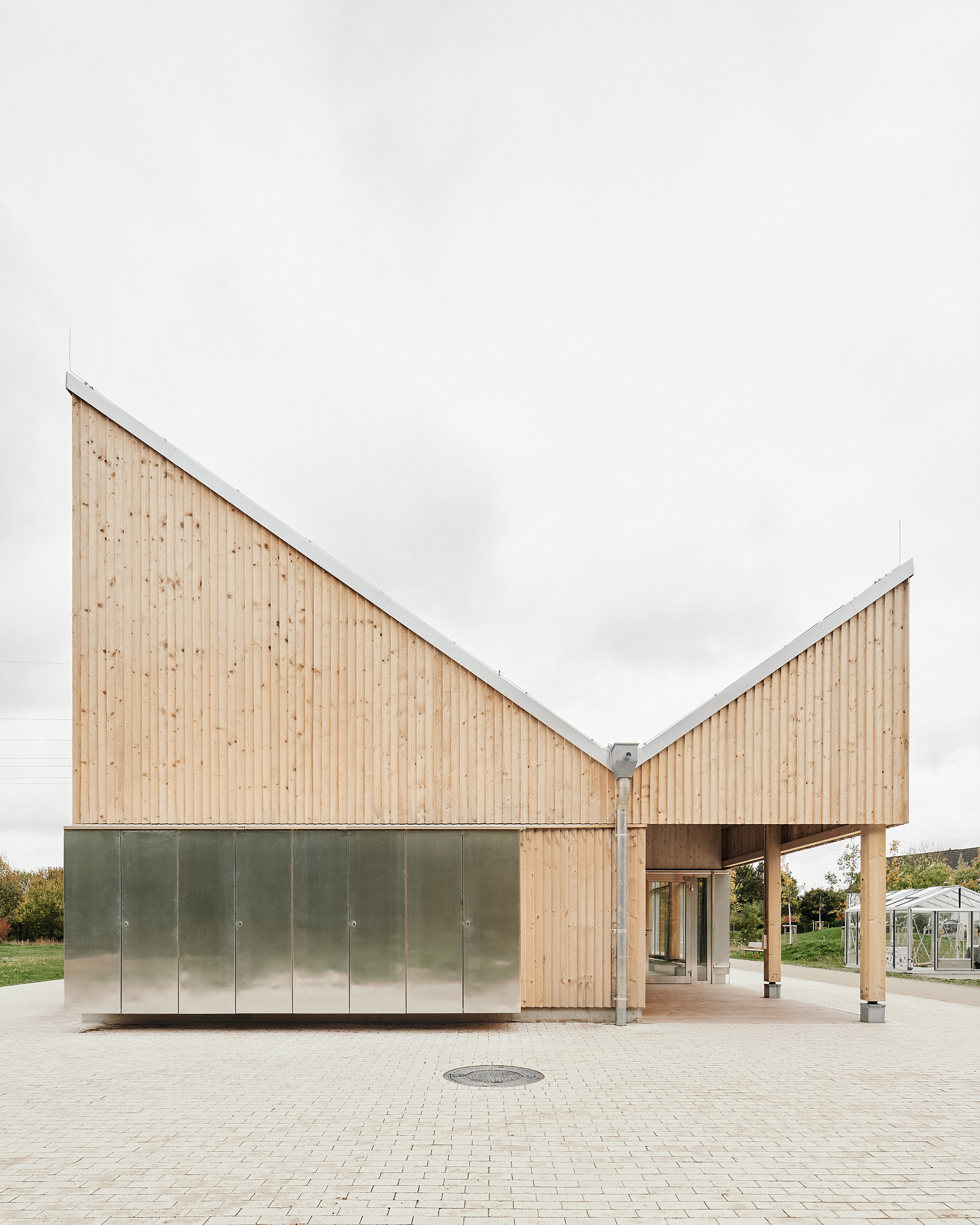 Close approach to the entrance from the brick-paved plaza, showing the folded zinc-clad base