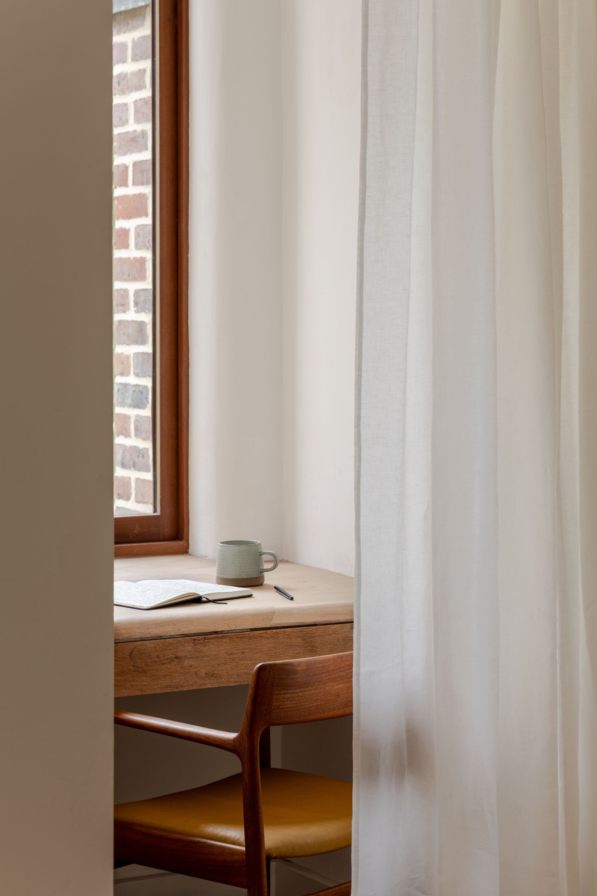 Detail of a writing desk with timber chair behind a sheer curtain beside a narrow window