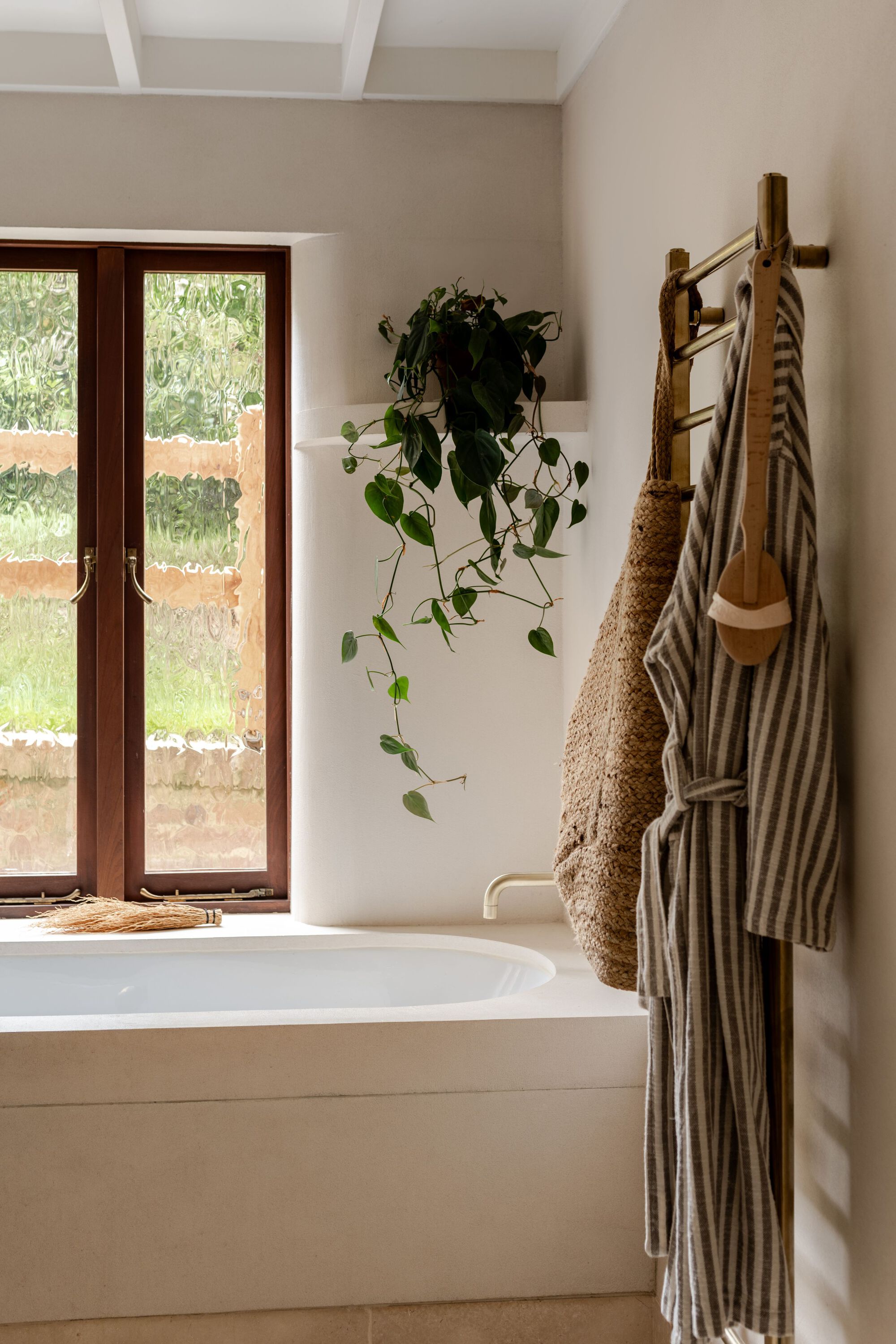 Bathtub with timber-framed window and hanging plant beside a ladder towel rail with striped linens