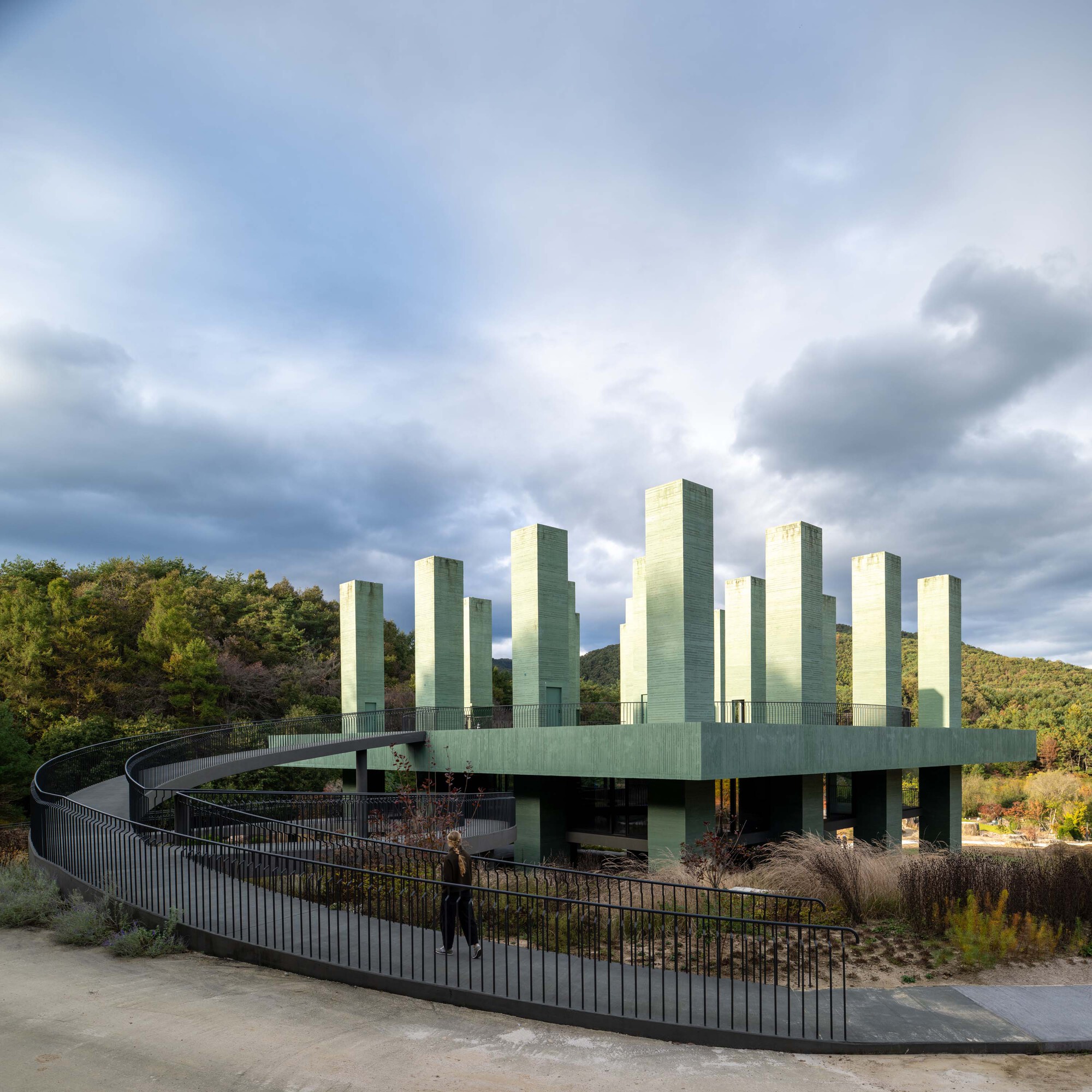 Elevated platform with vertical green concrete columns accessed by a curving ramp beneath dramatic clouds