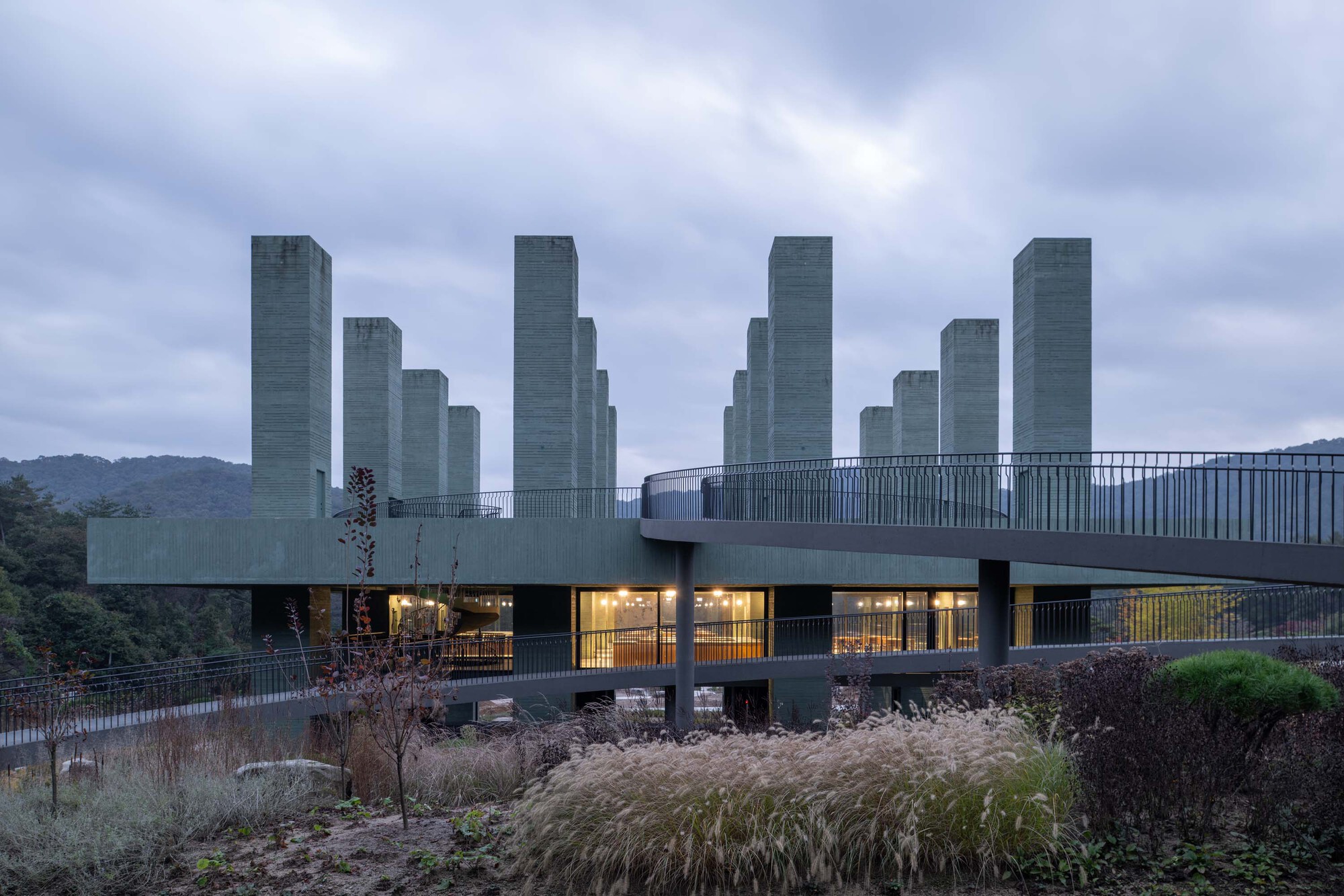 Elevated concrete structure with vertical fin columns above planted beds of ornamental grasses at dusk