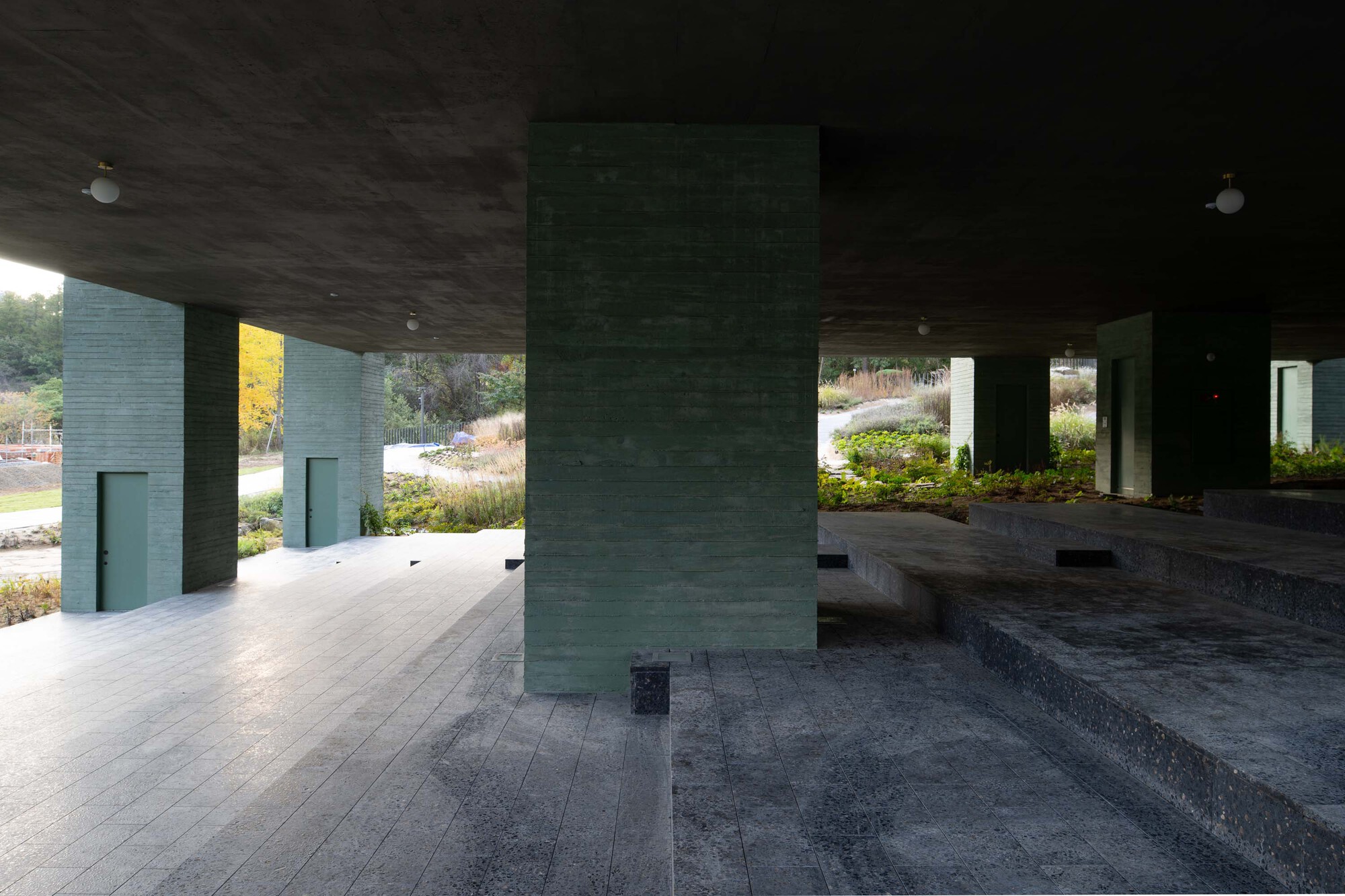 Sheltered plaza space with green concrete columns and terrazzo paving under a dark soffit