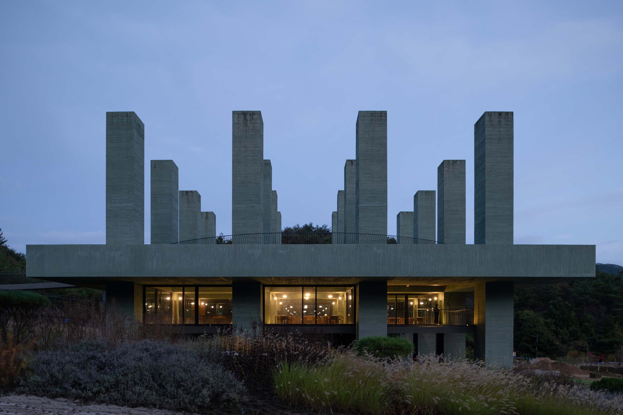 Elevated concrete slab with towers above a glazed base at dusk with interior lights glowing