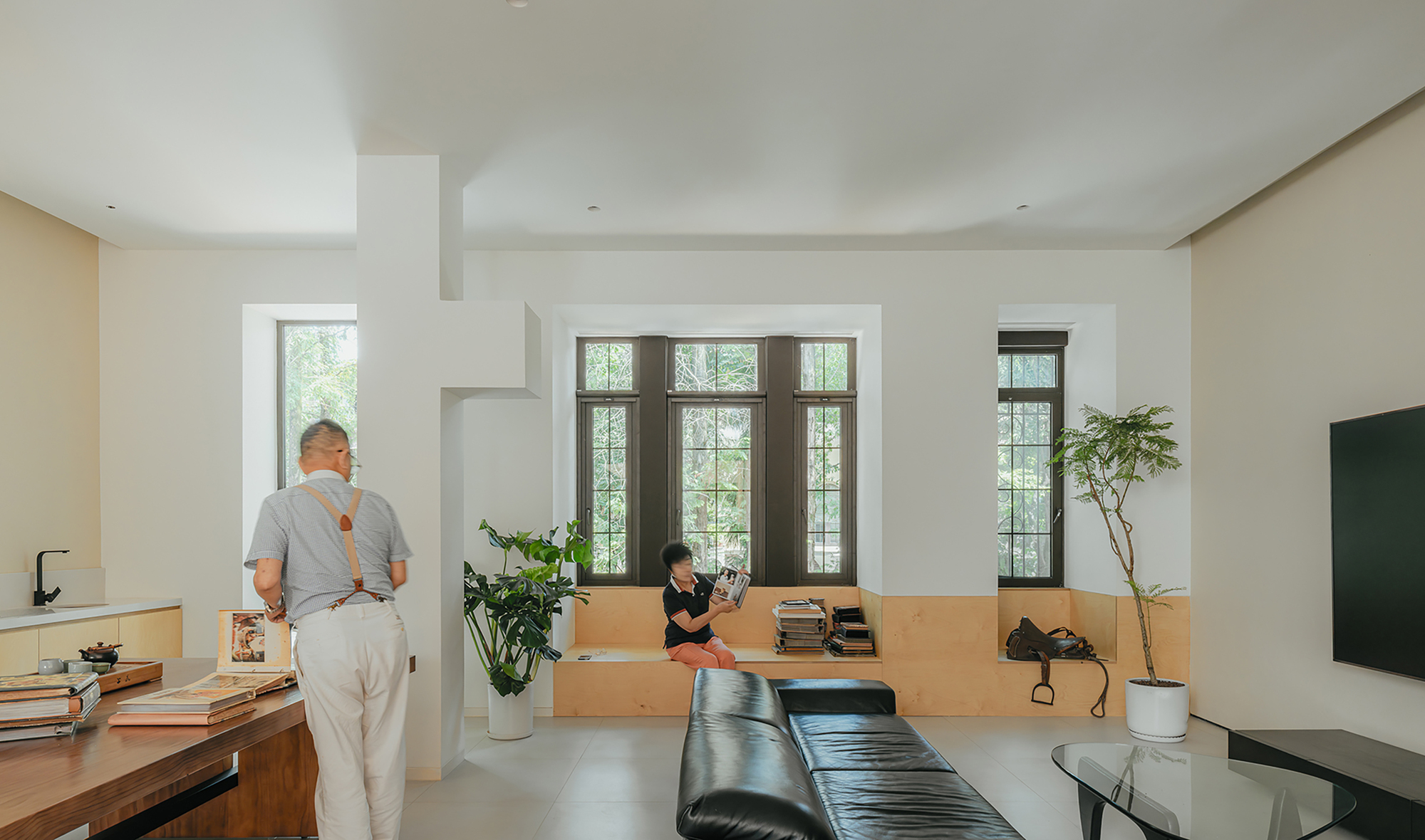 Open living space showing two people, built-in timber seating, and tall gridded windows overlooking greenery