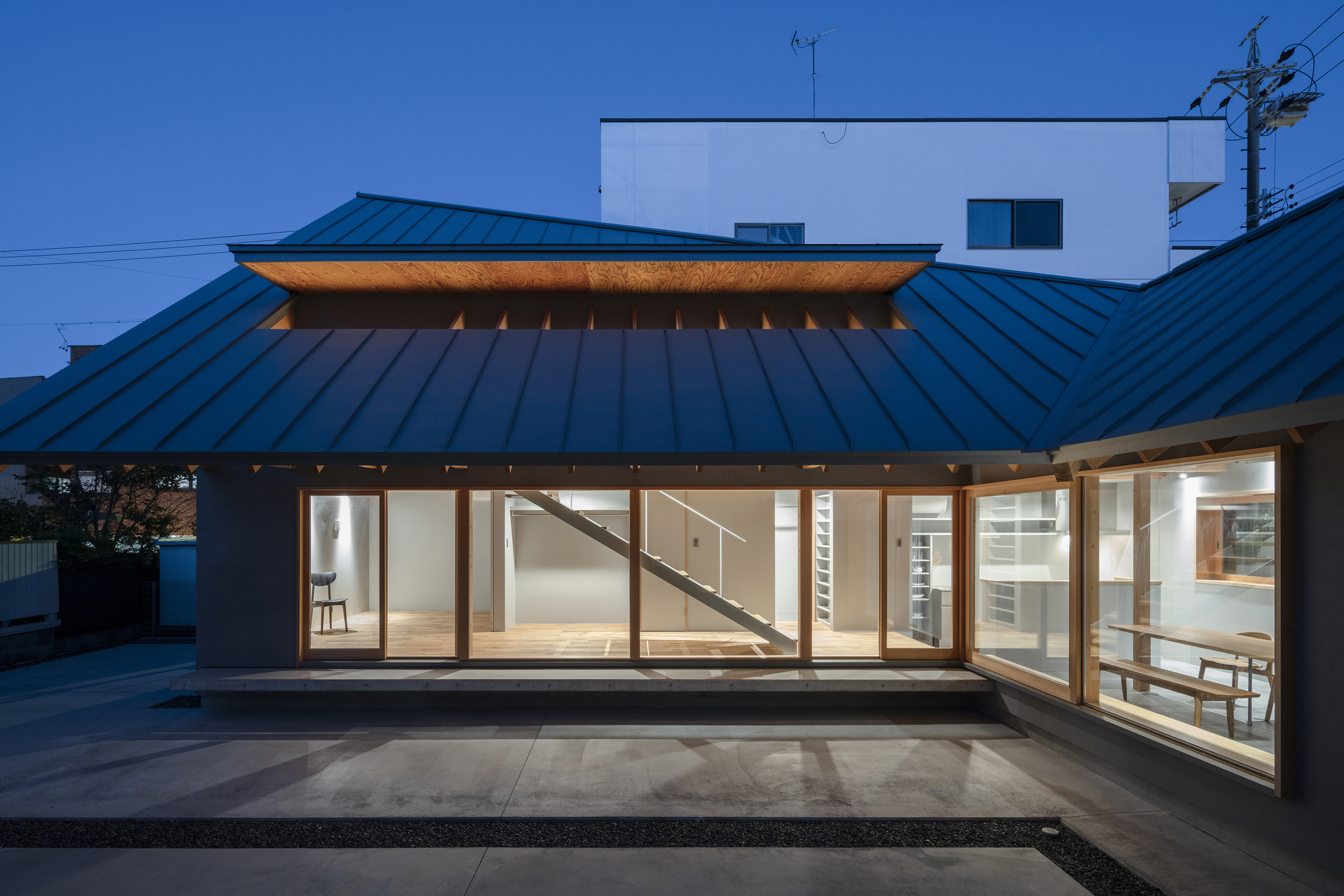 Evening view of the glazed facade beneath the metal roof with interior lighting and visible staircase