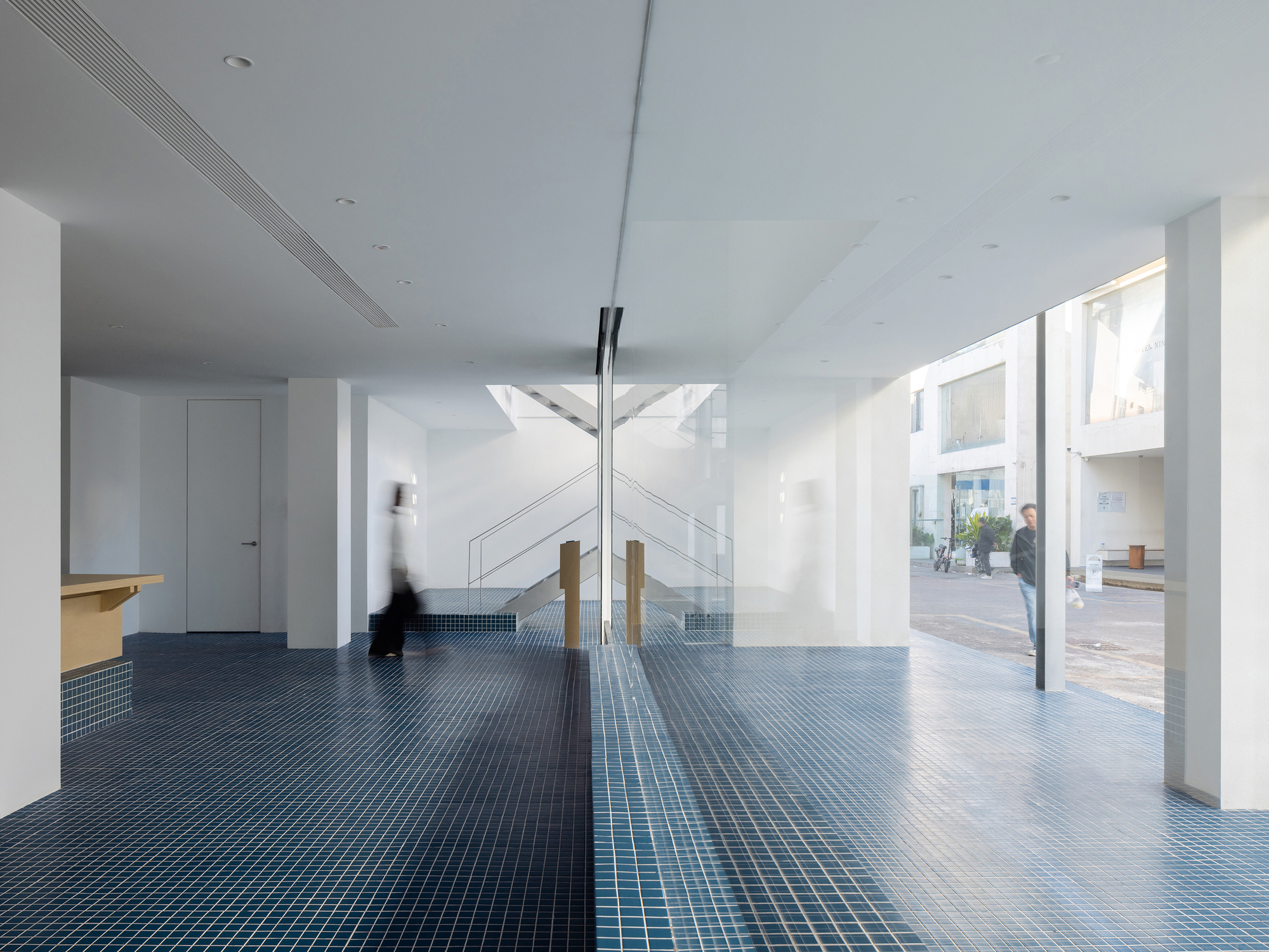 Double-height foyer with blue mosaic tile floor, timber columns, and figure passing through sunlit space