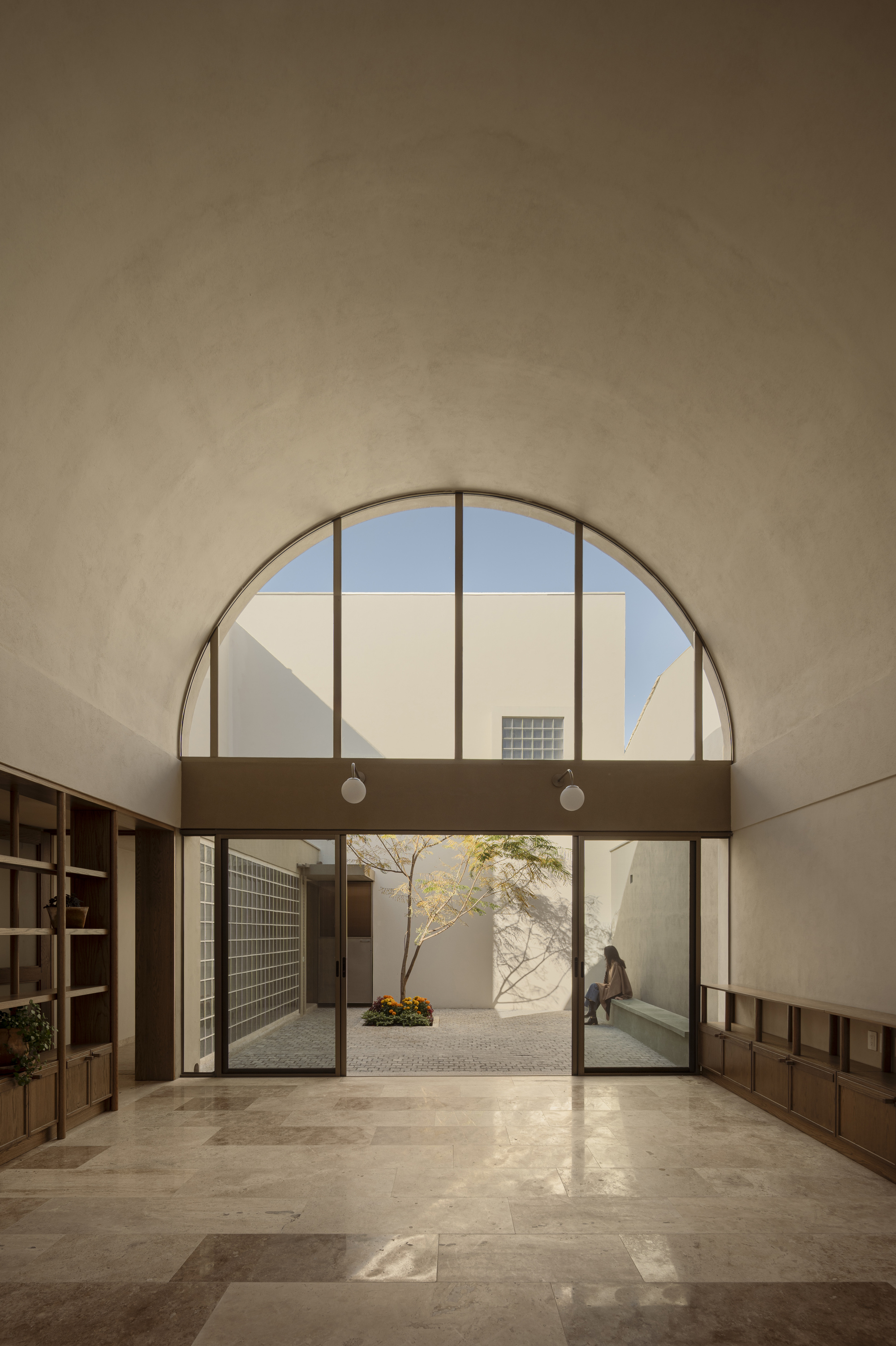 Arched window above glass doors opening to a sunlit courtyard with a figure passing through