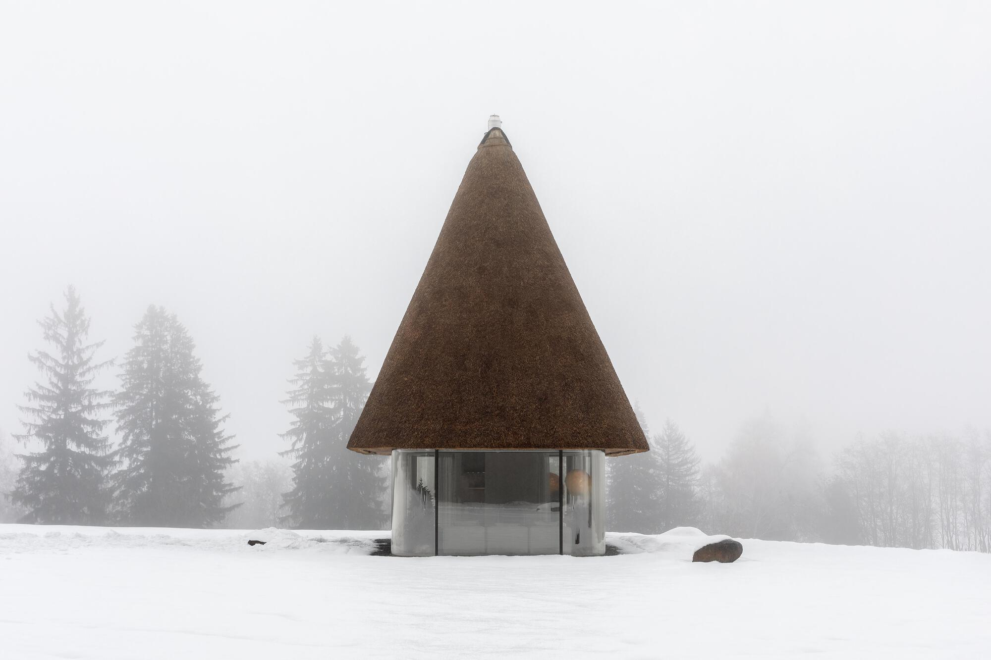 Thatched pyramidal roof pavilion standing in a snow-covered field surrounded by misty pine trees