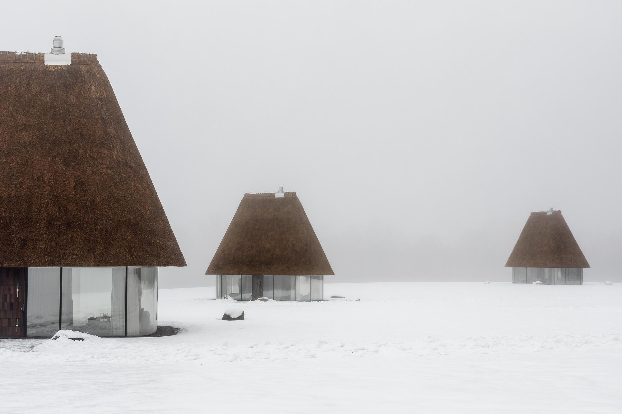 Three thatched roof pavilions of varying sizes dispersed across a white snowfield in heavy fog