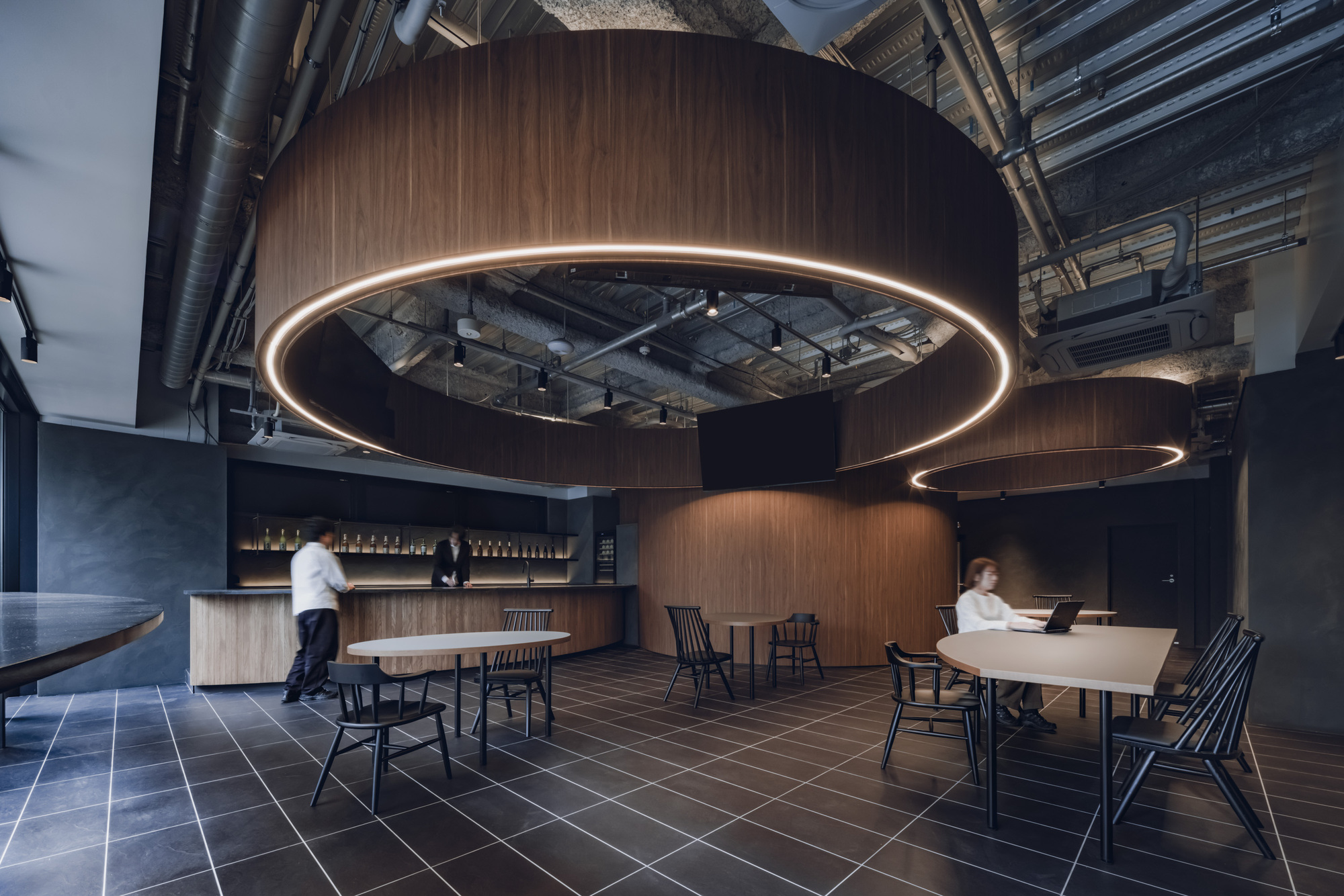 Dining area with curved wooden pods and illuminated ring fixtures as two figures move through the space