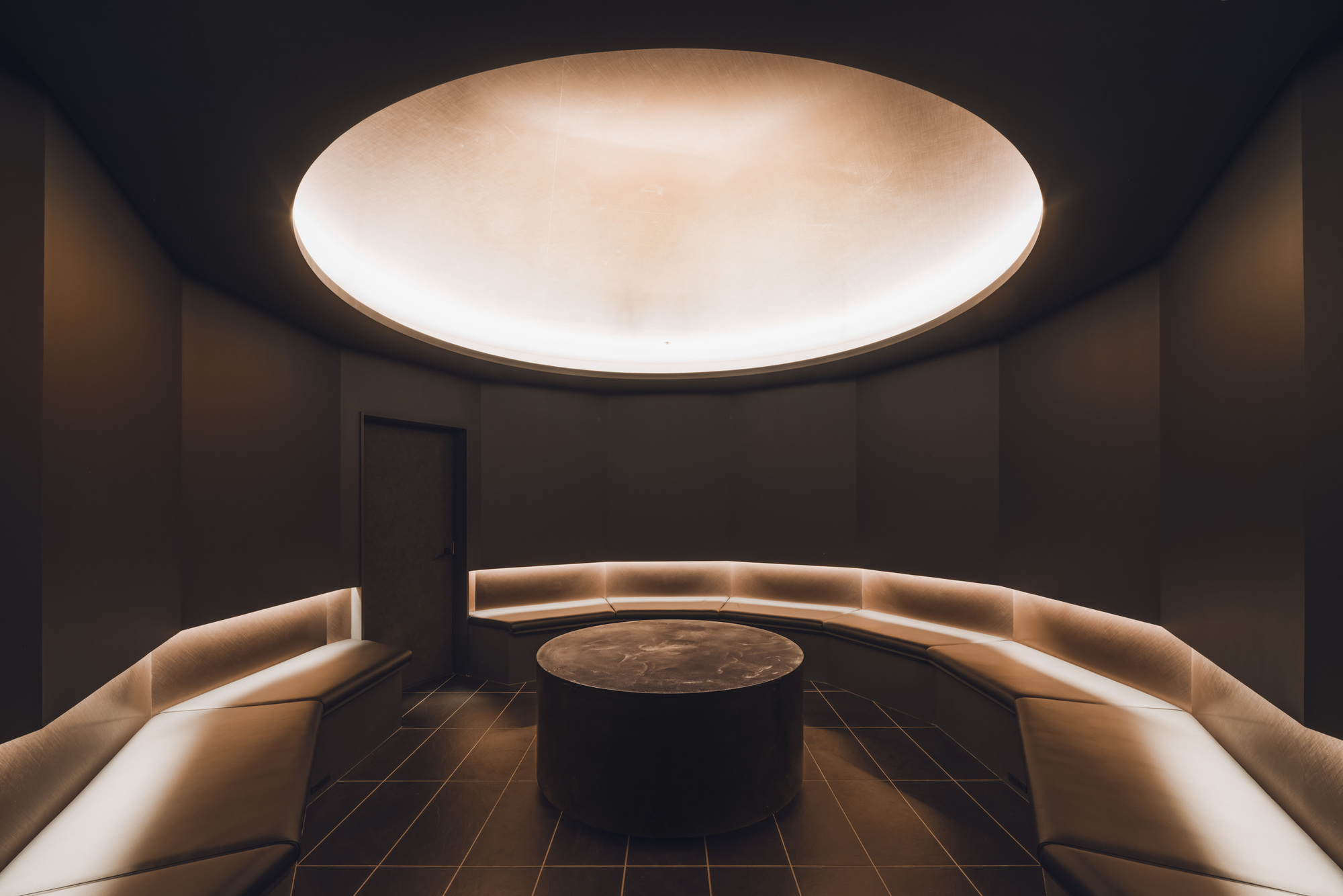 Circular room with backlit curved banquettes surrounding a central table beneath an illuminated ceiling oculus