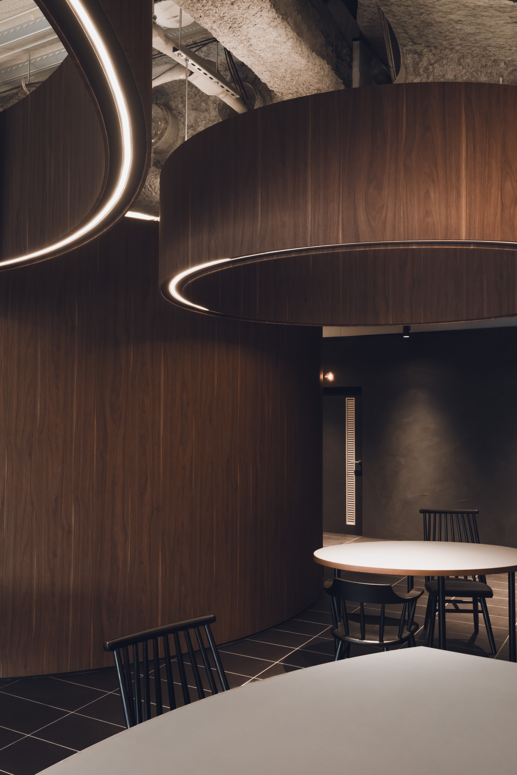 Dining area with circular walnut veneer volumes and illuminated ring details suspended from an exposed concrete ceiling