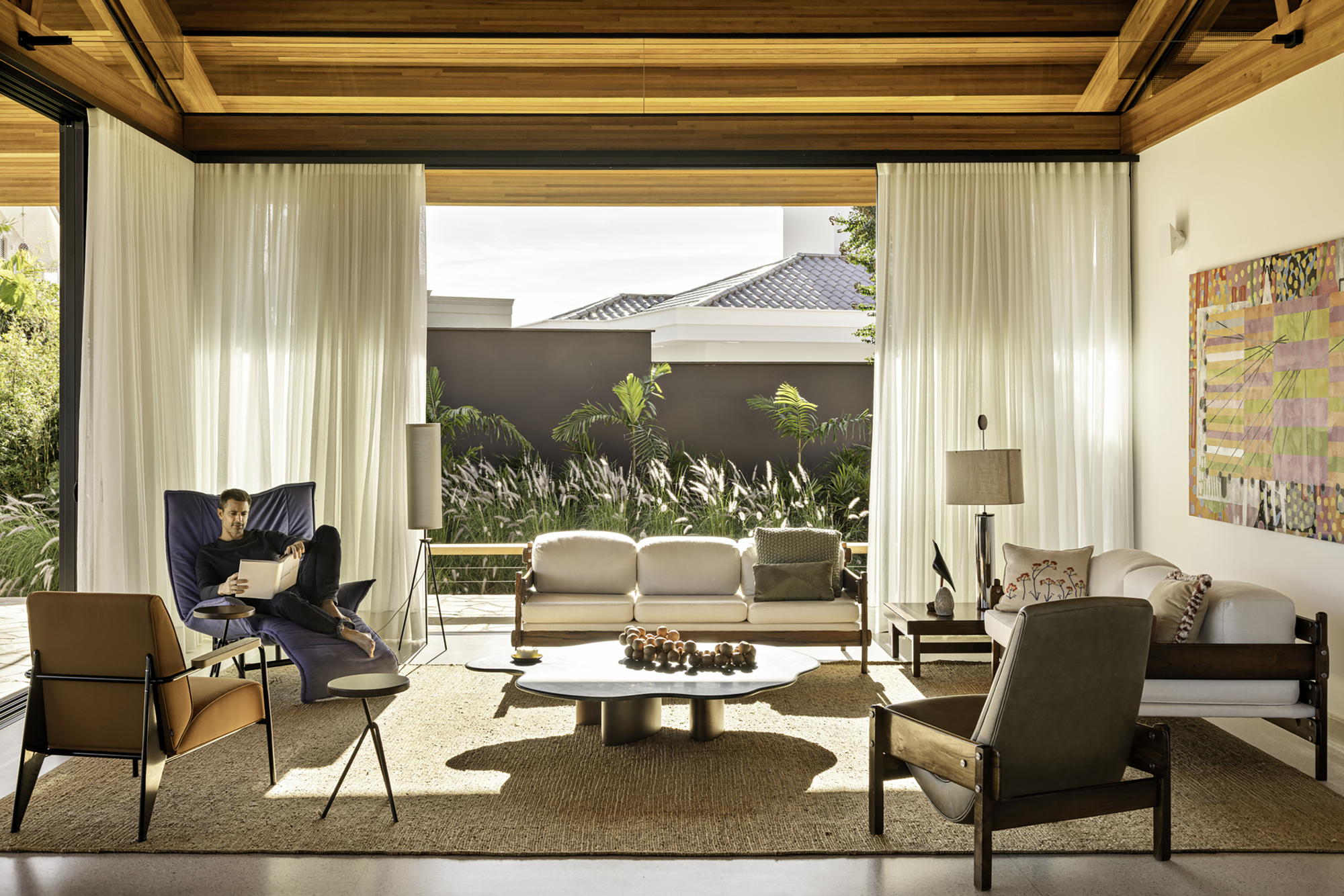 Living room framed by floor-to-ceiling curtains opening to a planted courtyard with a seated figure reading