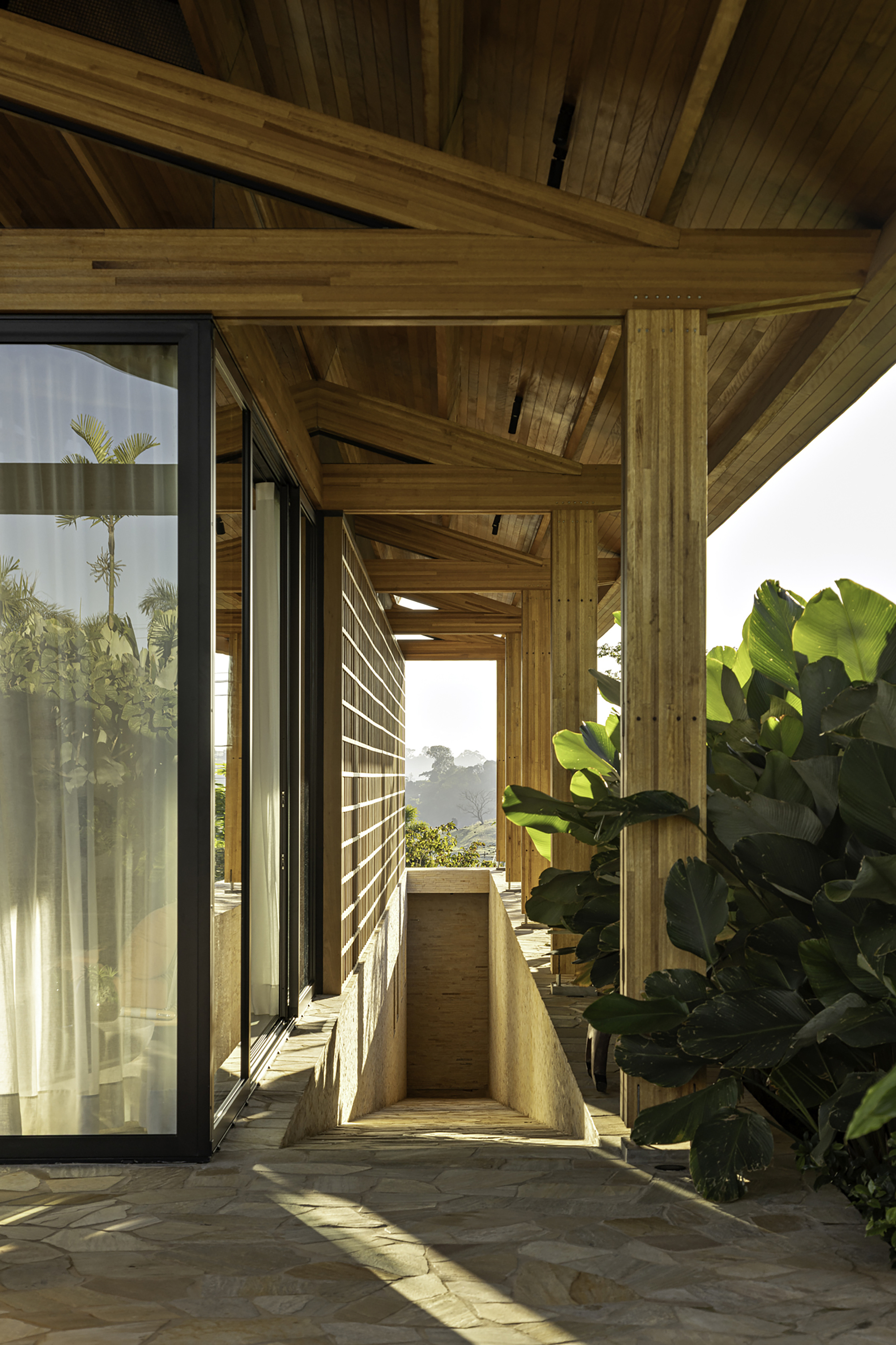 Covered walkway with timber columns and horizontal slatted screen framed by banana leaves