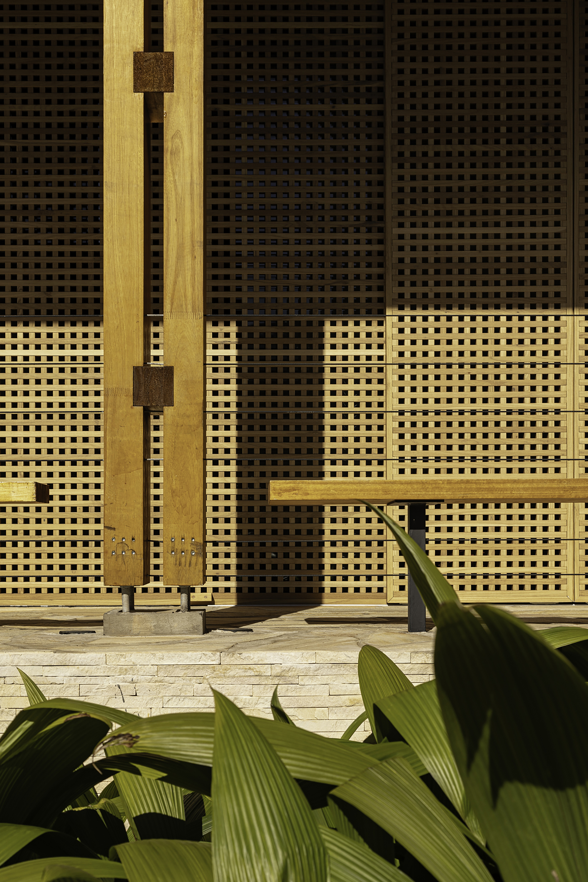 Close-up of woven lattice facade panels in shadow with timber post and tropical plant leaves below