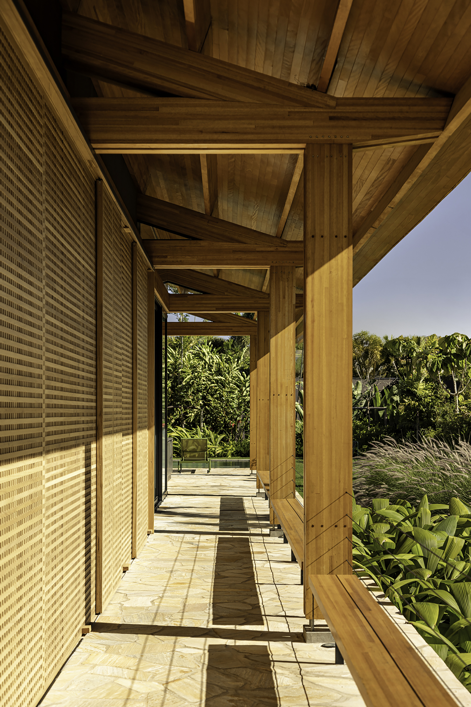 Covered timber walkway with woven screen wall casting dappled shadows on sandstone pavers at midday