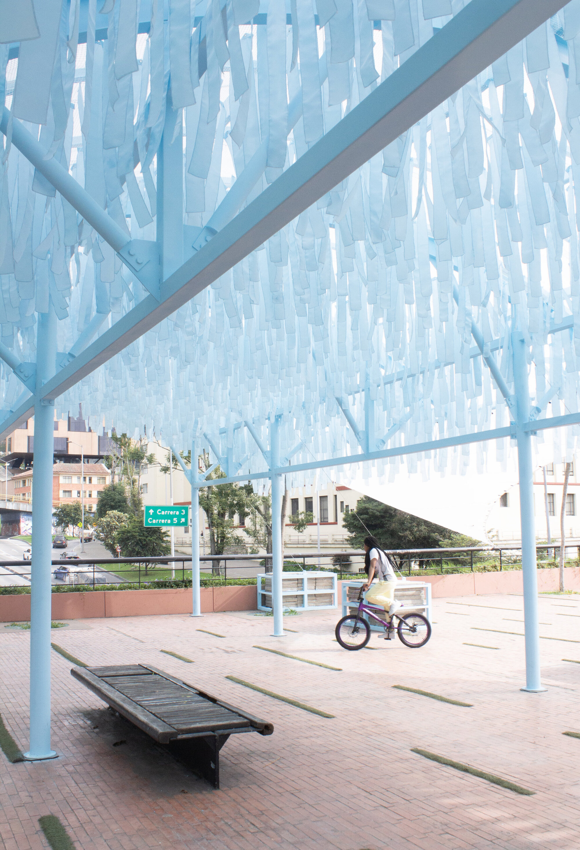 Underside of the translucent vertical fin canopy with steel supports above a plaza bench and bicycle