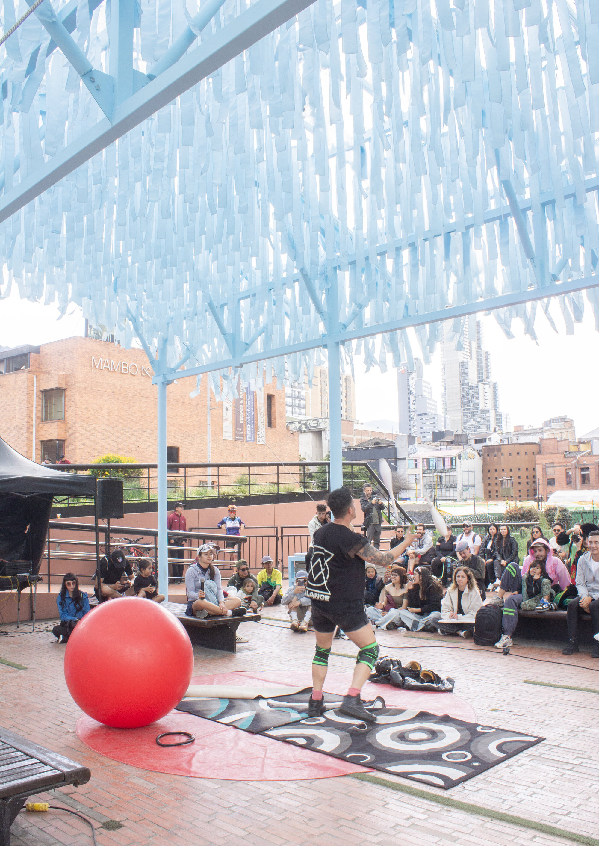 Translucent blue canopy overhead sheltering a public performance space with seated audience and large red ball