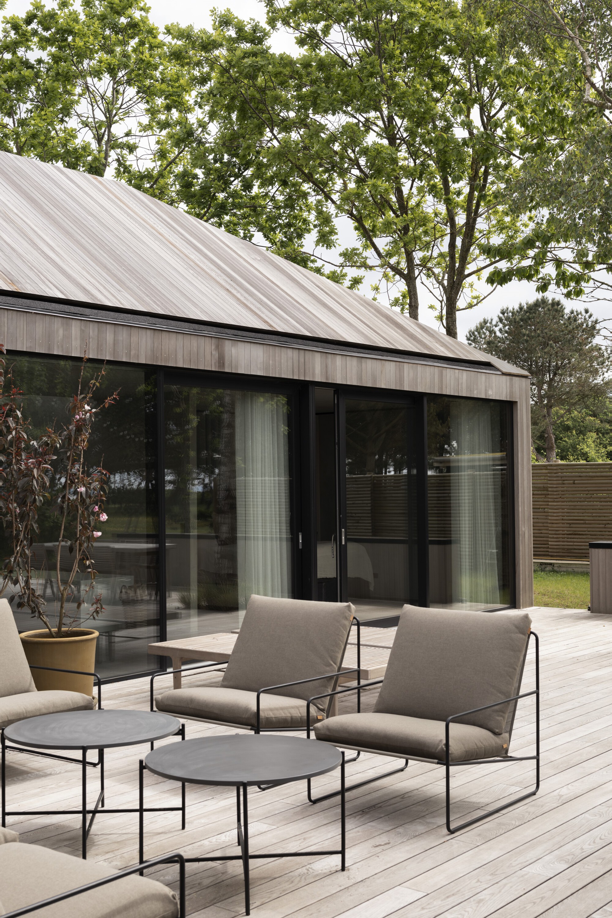 Timber deck with metal-framed seating beneath a sloped metal roof and surrounding deciduous trees