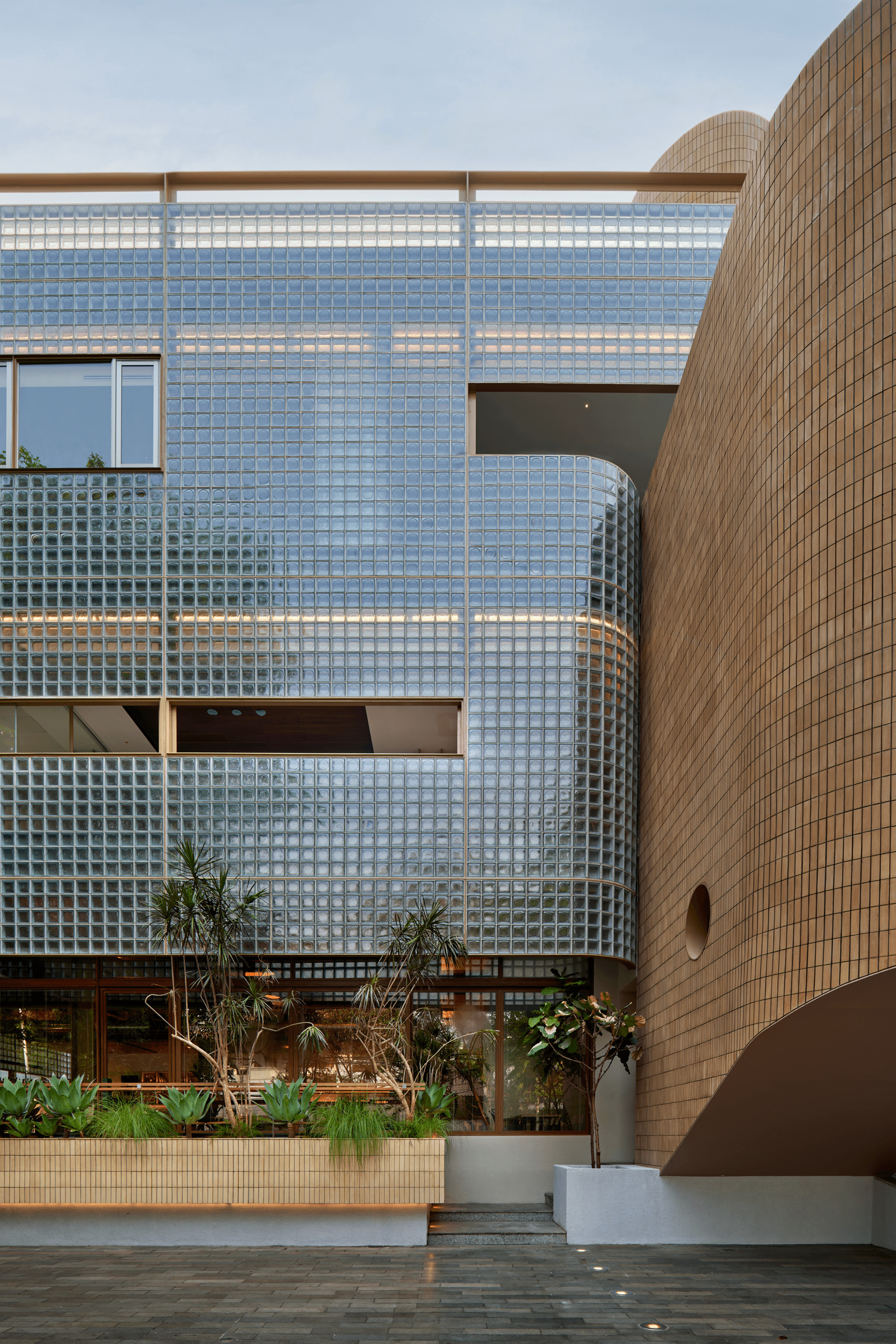 Glass block wall with rectangular openings beside cylindrical brick tower and ground level planted beds