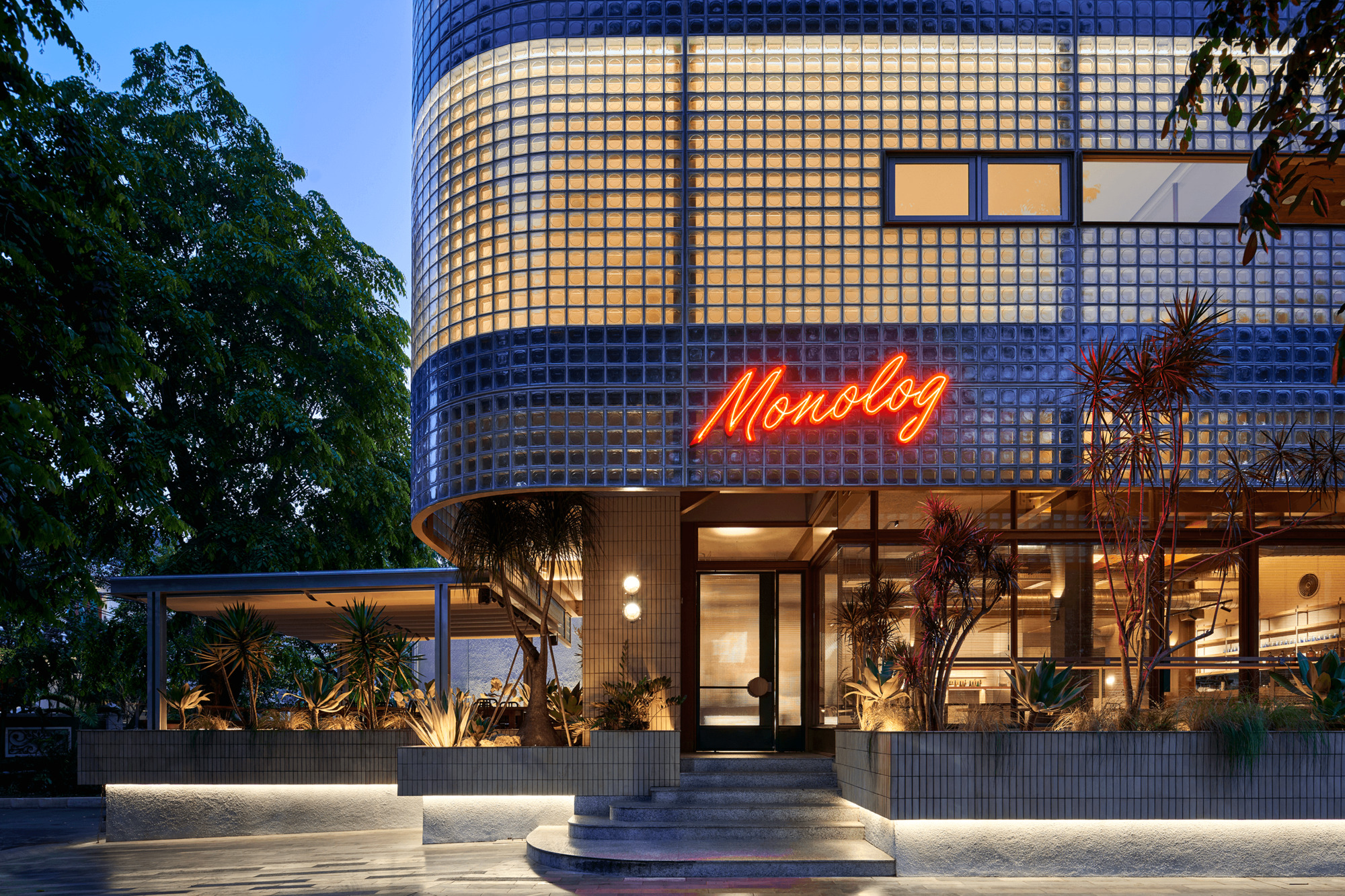 Entrance with red neon signage above glass doors and planted terrace at dusk