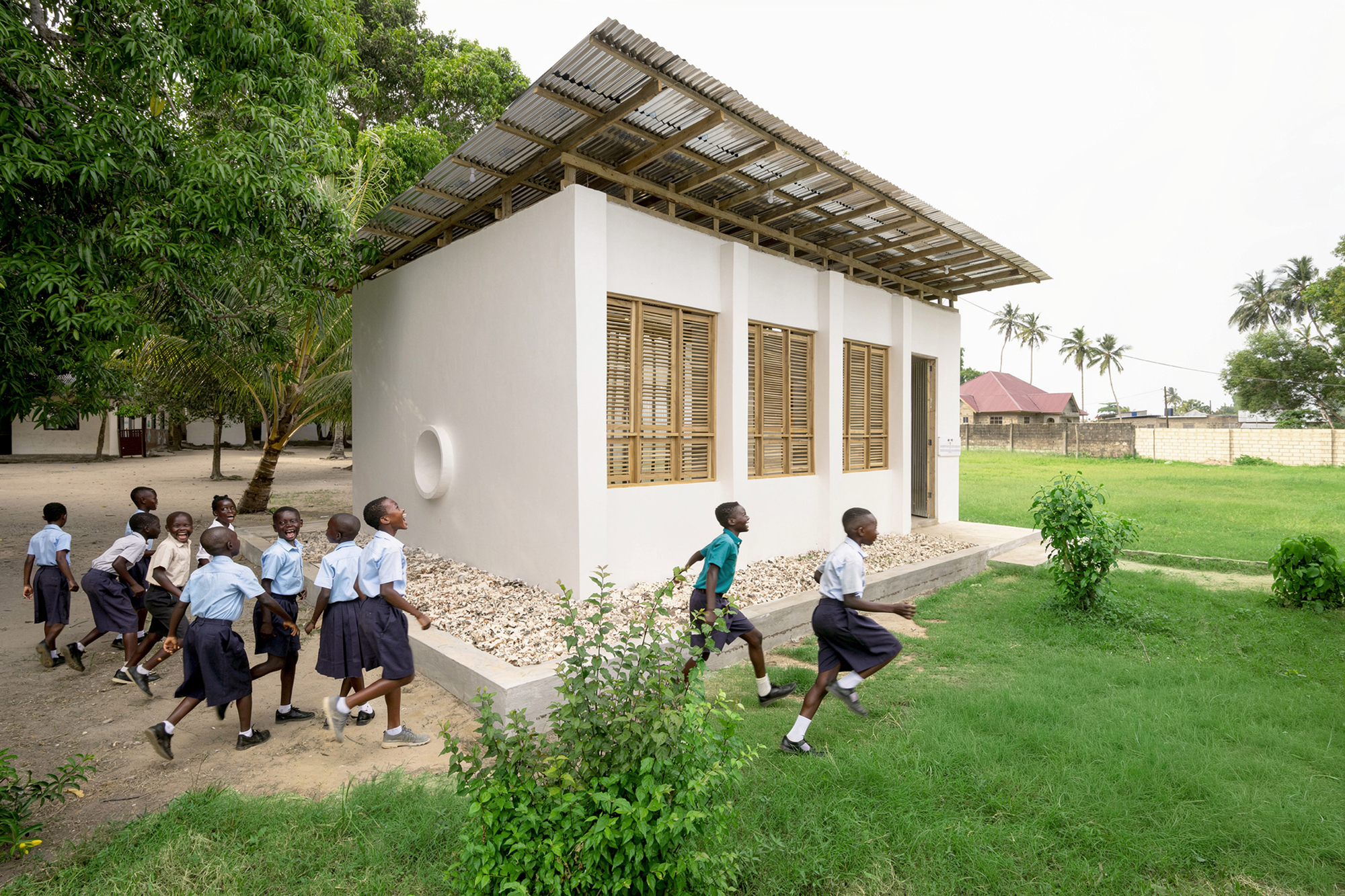 White plastered classroom with louvered timber windows and corrugated metal canopy as children run past