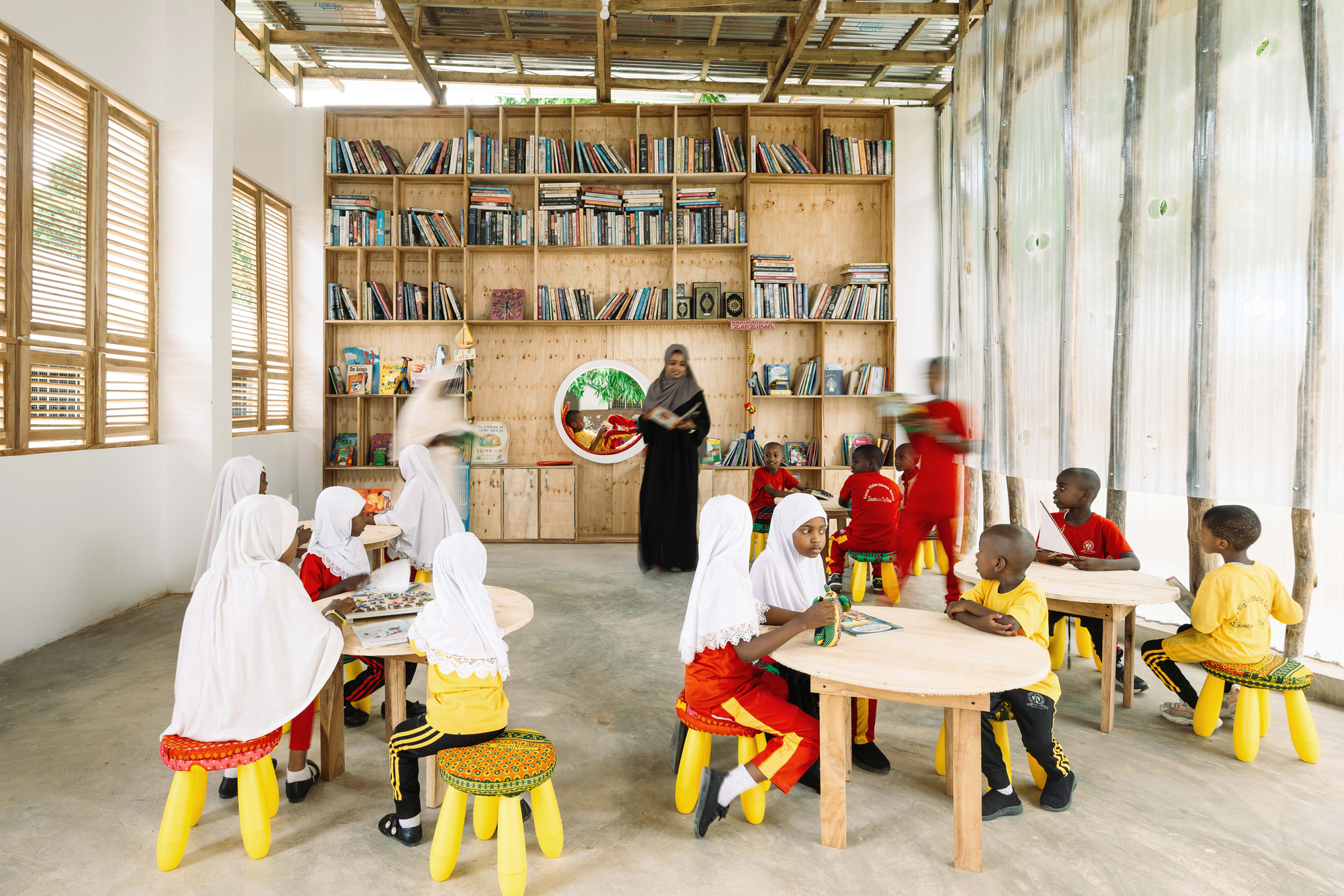 Children seated at low tables beneath exposed timber roof trusses with teacher standing by shelving wall