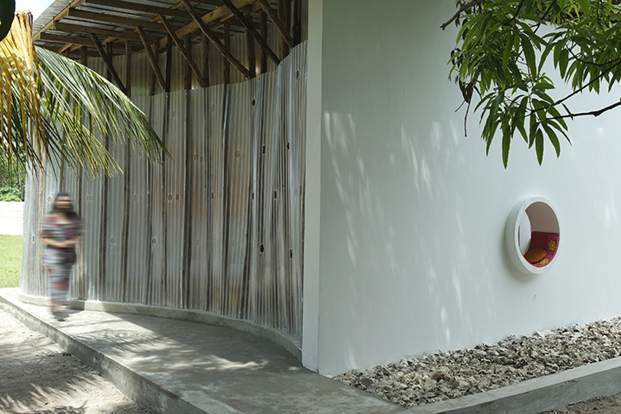 Covered walkway with vertical timber screens and a circular window opening as a figure passes through dappled light