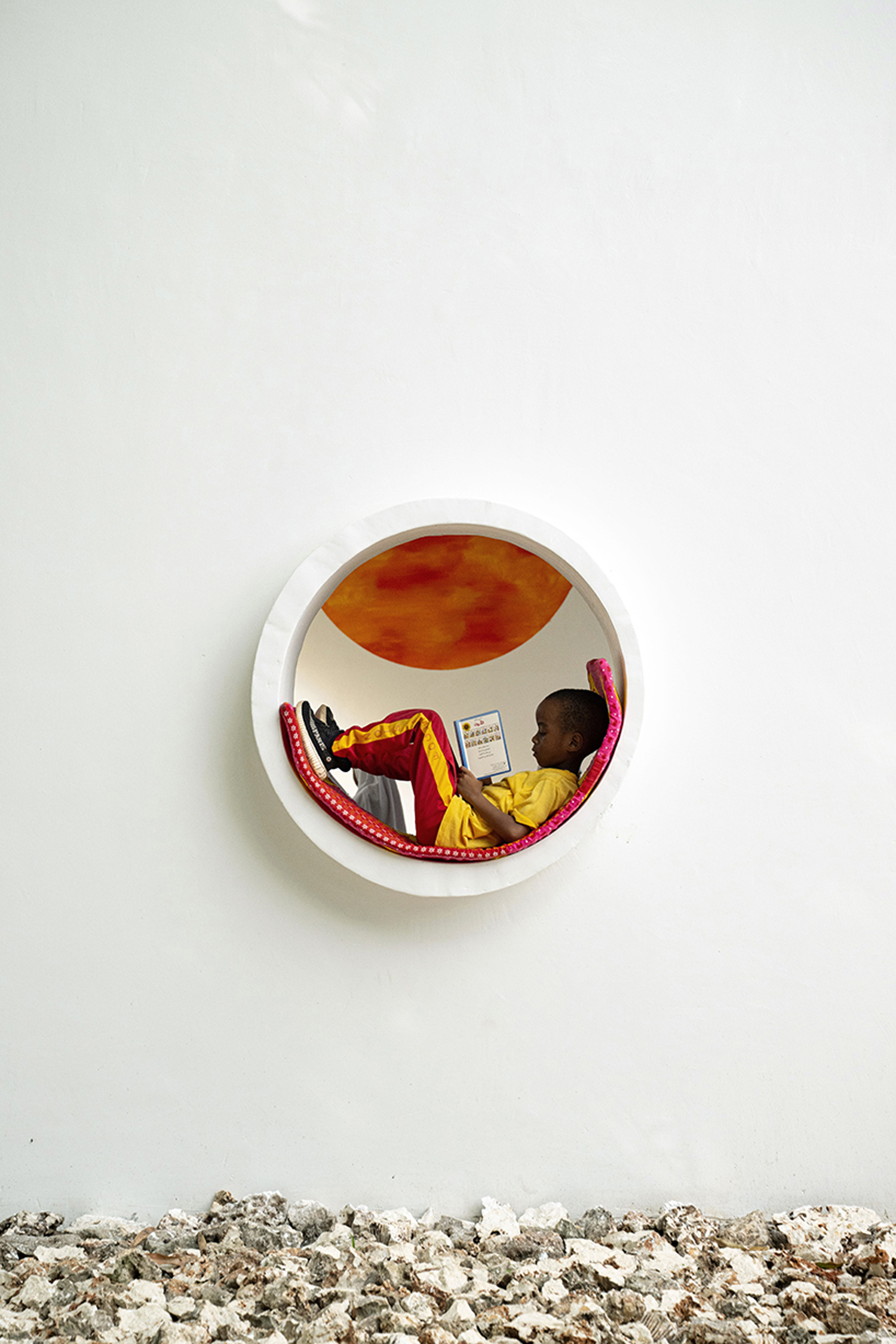 Circular opening in white wall revealing a child reading inside a nook with orange-painted interior and gravel below