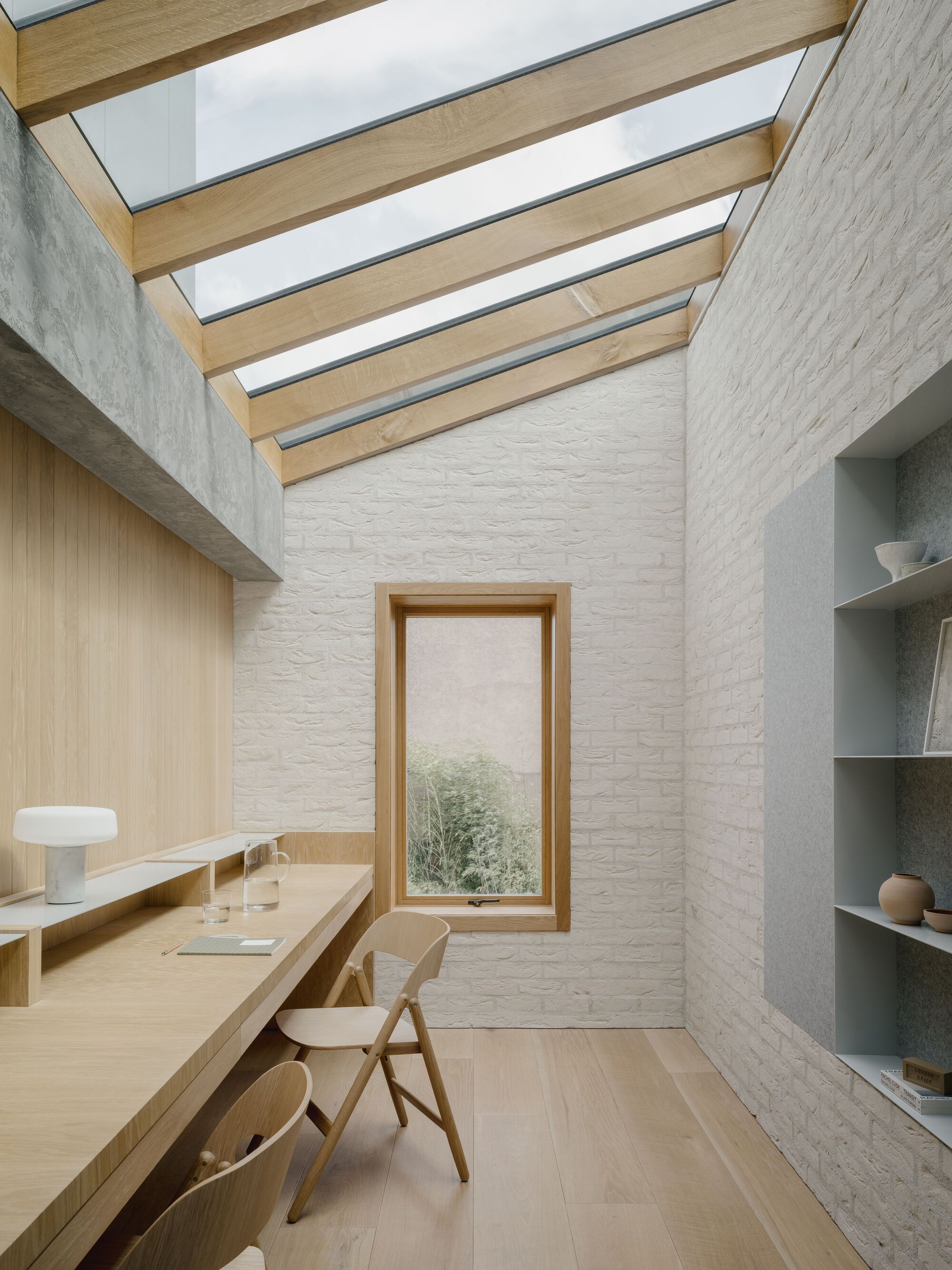 Study with timber beam skylight strips above white brick walls and vertical window overlooking greenery