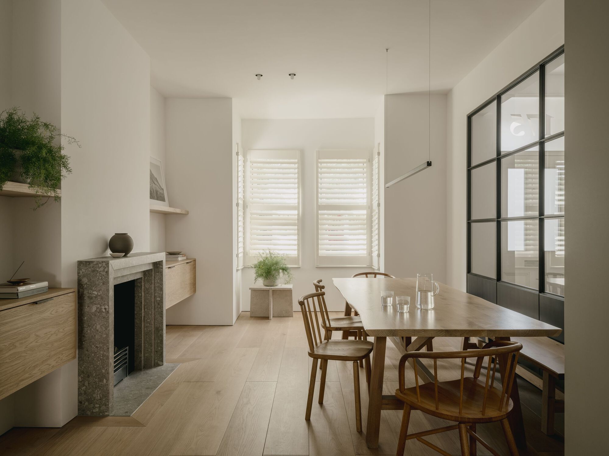 Dining area with oak table and chairs beside a steel-framed glass screen and louvered shutters