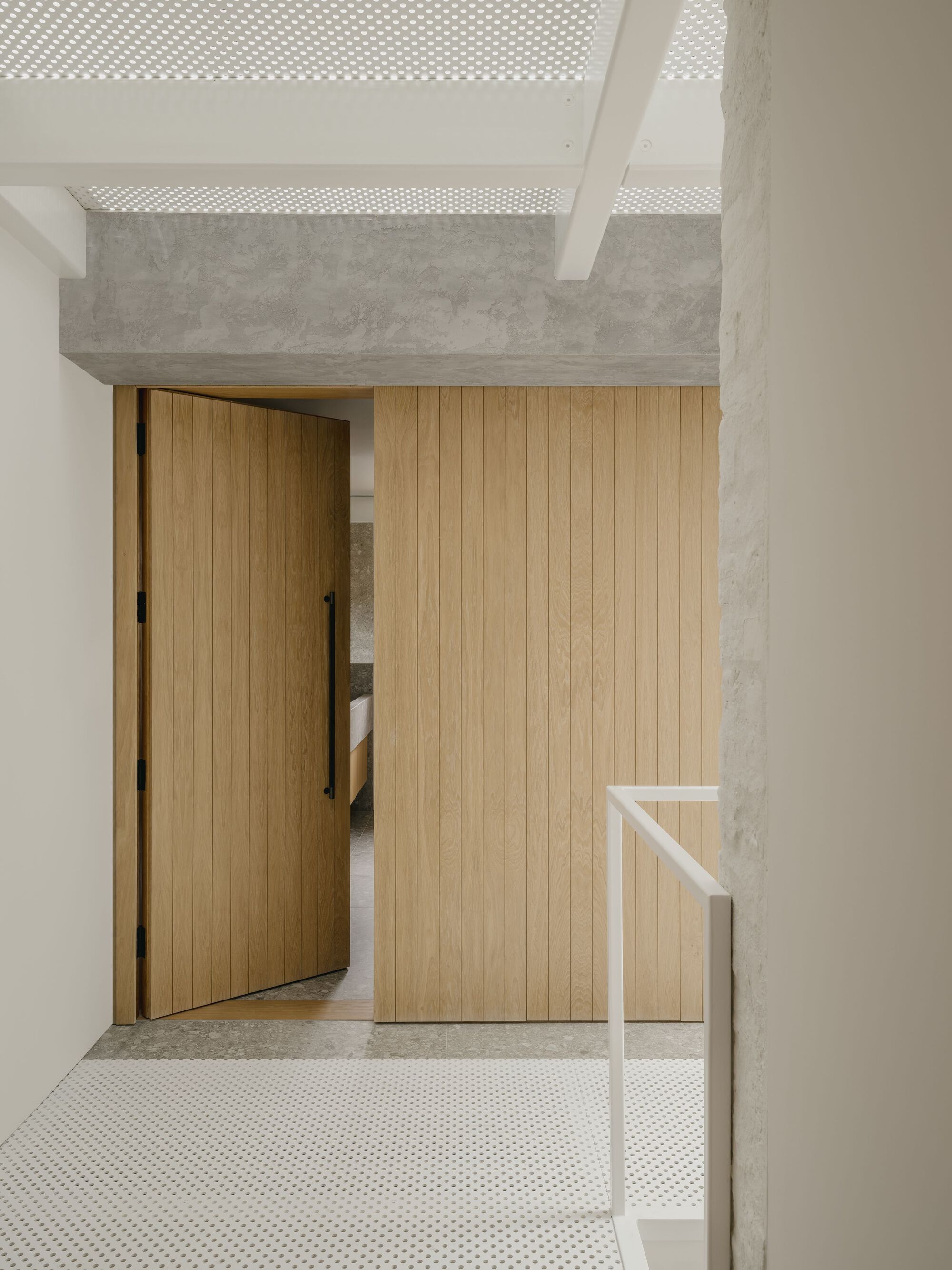 Timber sliding door and slatted wall panel beneath a perforated concrete ceiling beam with dappled light
