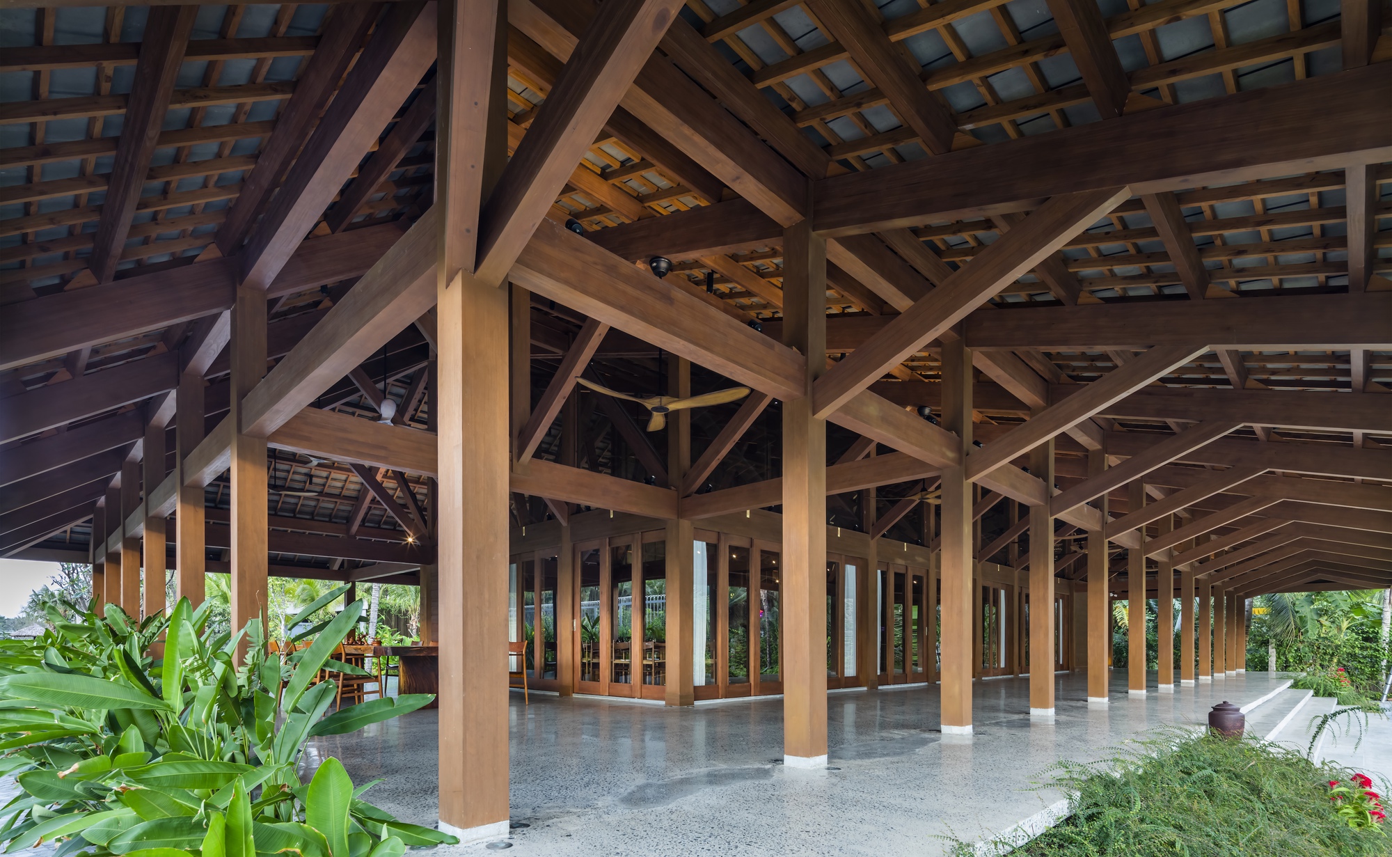 Open-sided pavilion with exposed timber trusses and columns framed by tropical plantings