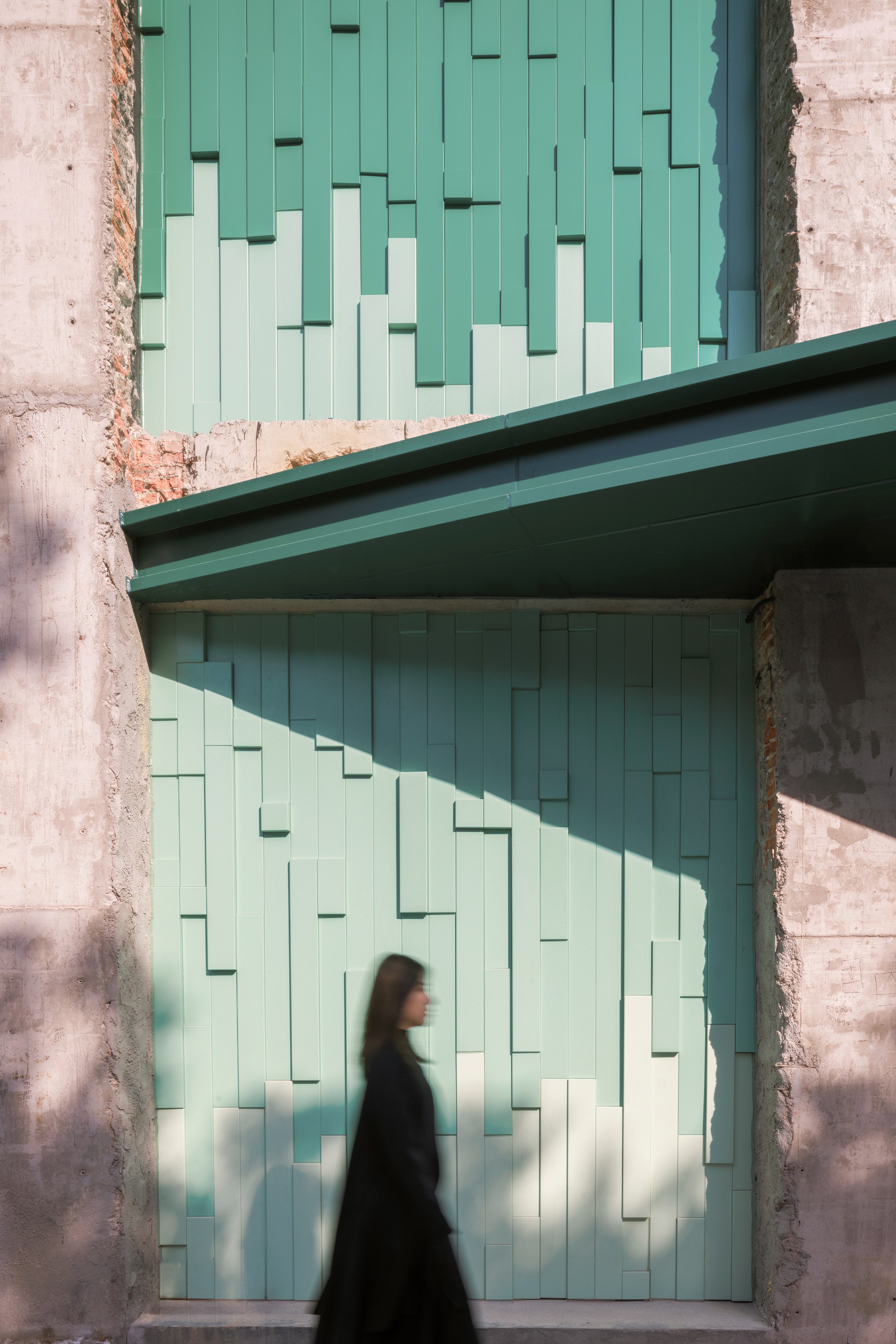 Blurred figure passing beneath a pixelated green tile wall within a rough concrete opening