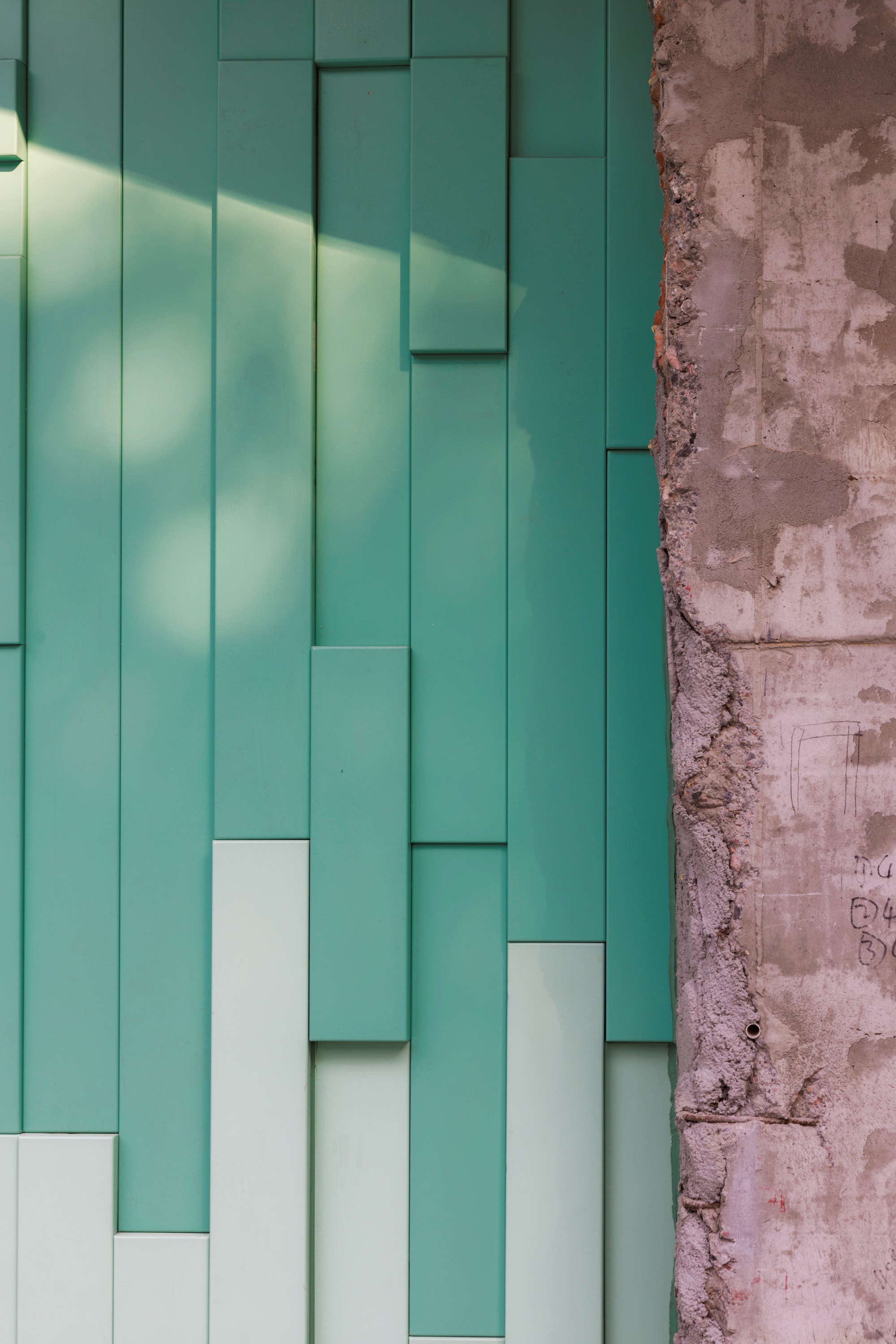 Staggered mint green metal panel facade meeting a weathered pink concrete wall in sunlight