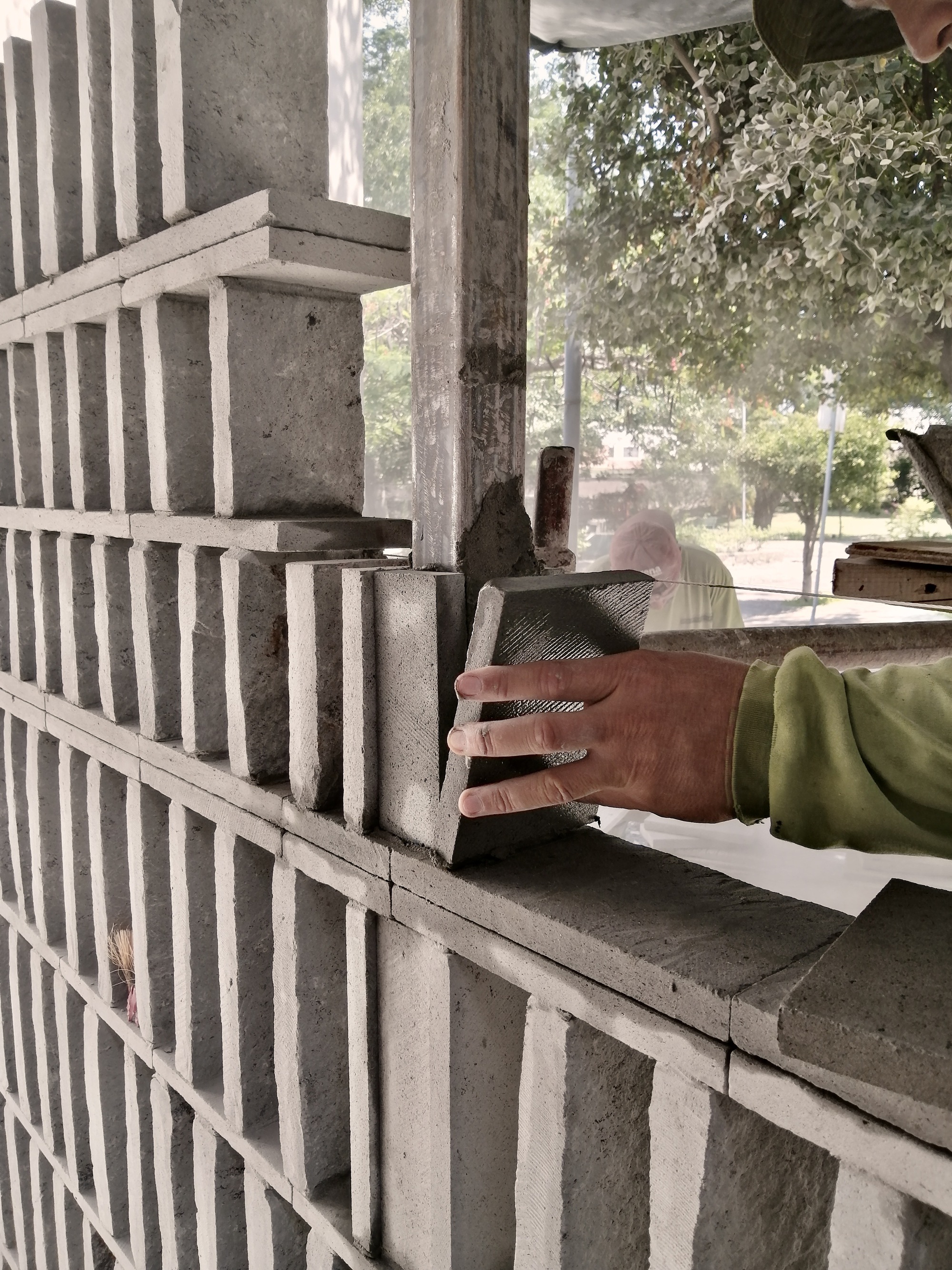 Hand placing custom concrete block into perforated masonry wall during construction with trees visible beyond