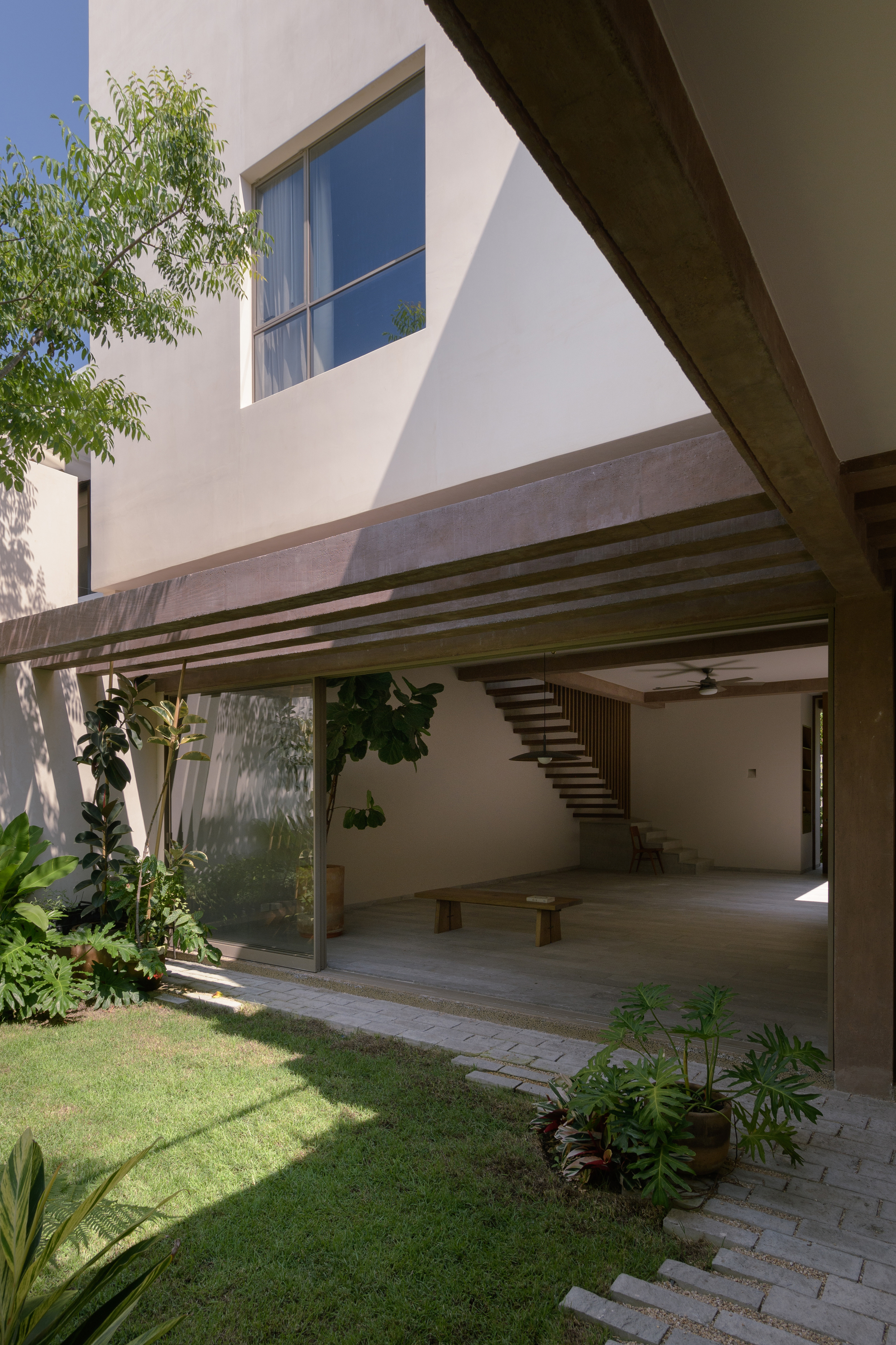 Covered terrace with exposed concrete beams and open staircase framing a planted courtyard under blue sky