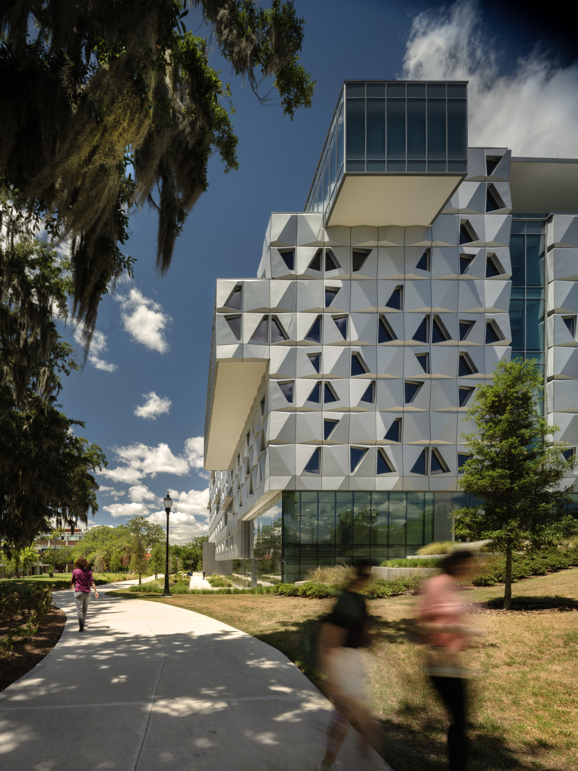 Angled view of the geometric panel facade and glazed base with pedestrians walking along the shaded path