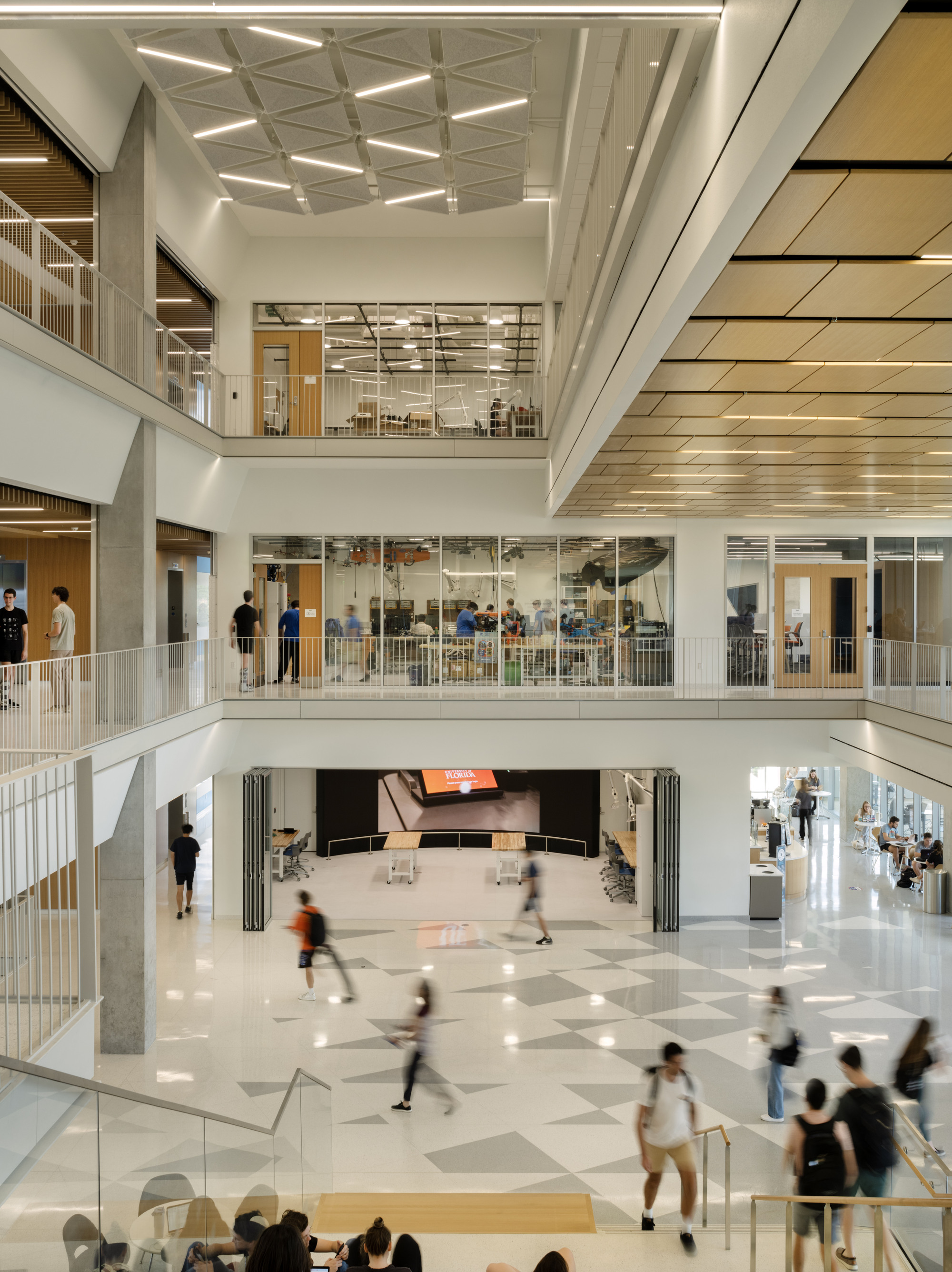 Multi-story atrium with stepped wooden seating and people moving across the white floor below