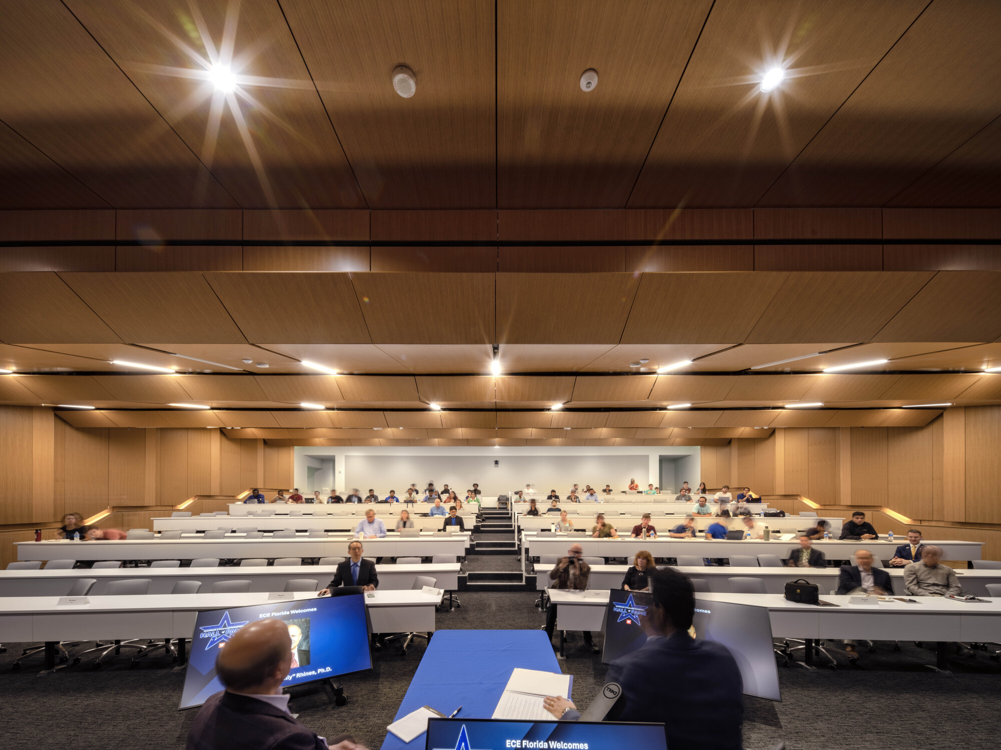 Tiered lecture hall with undulating wood ceiling panels and recessed strip lights illuminating rows of students