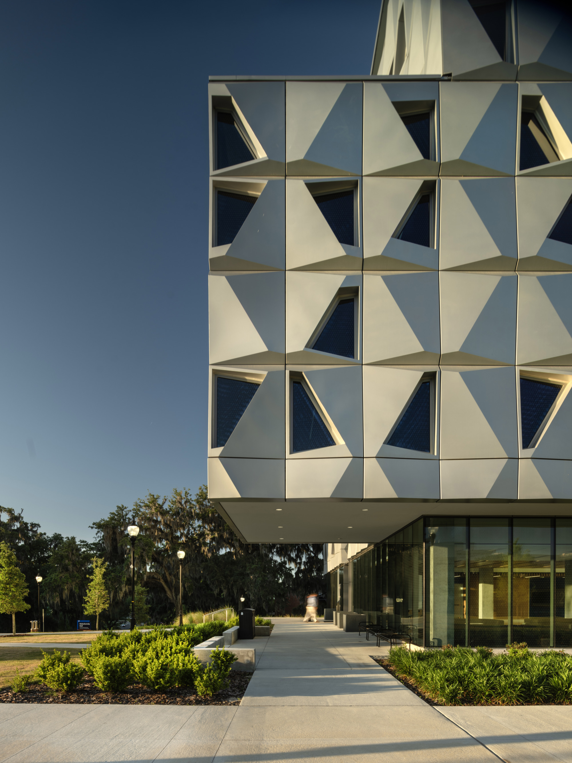 Geometric folded panel facade above glass-walled ground floor and landscaped entry plaza at dusk