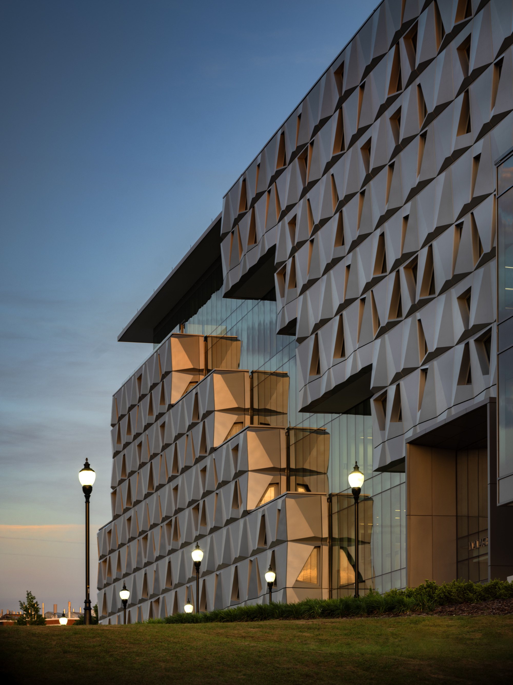 Angular facade with illuminated corner stair tower and street lamps along the sloped lawn at twilight