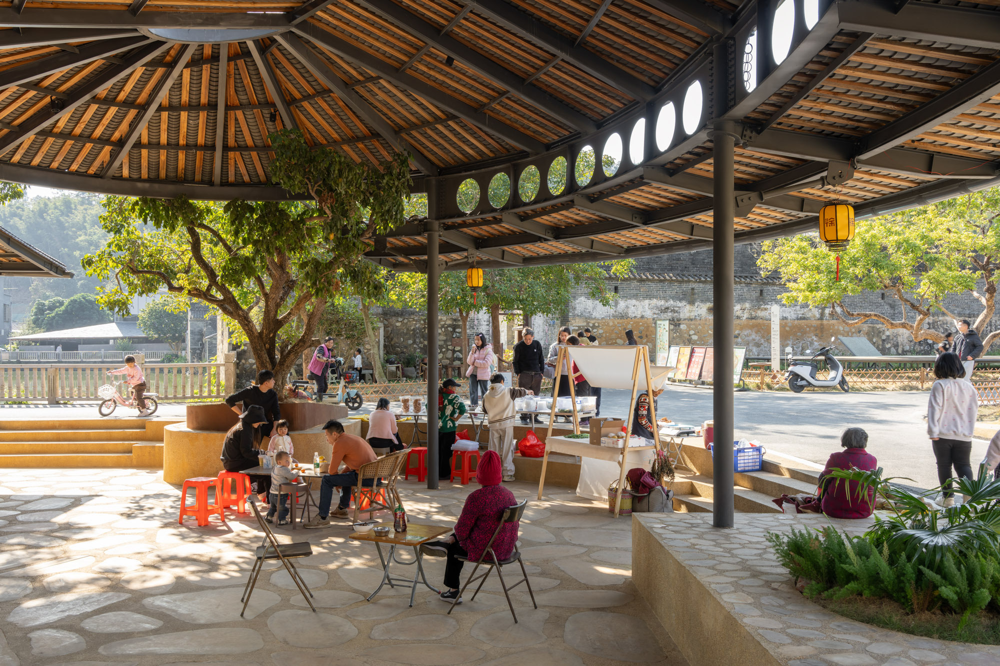 Open timber pavilion with bamboo ceiling and circular openings sheltering children engaged in activities on flagstone paving