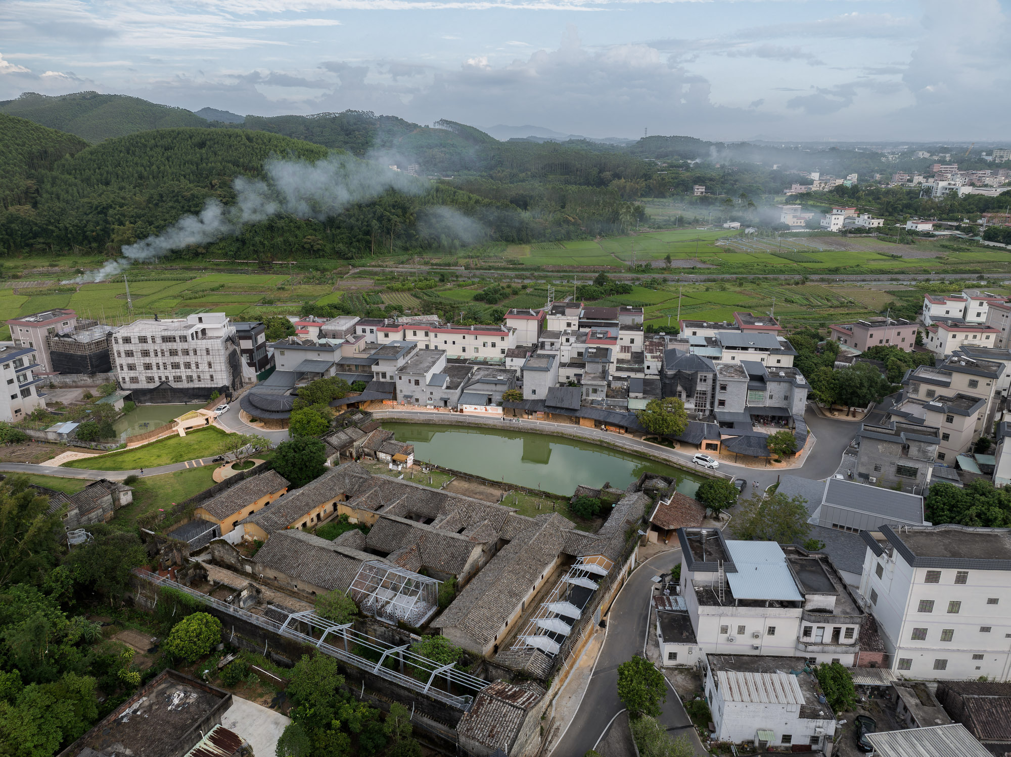 Aerial view of a village with tiled roofs surrounding a central pond and green fields in mist