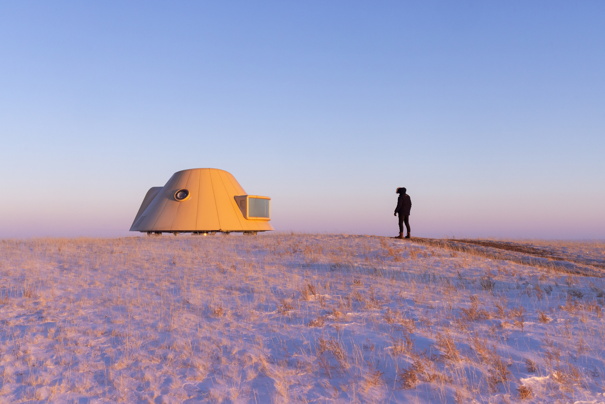 Person standing beside dome-shaped shelter on a snow-covered ridge in golden sunset light