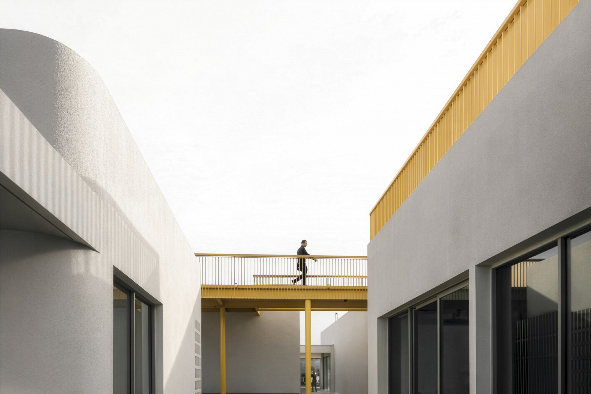 Elevated bridge with yellow railing connecting two white volumes with person crossing